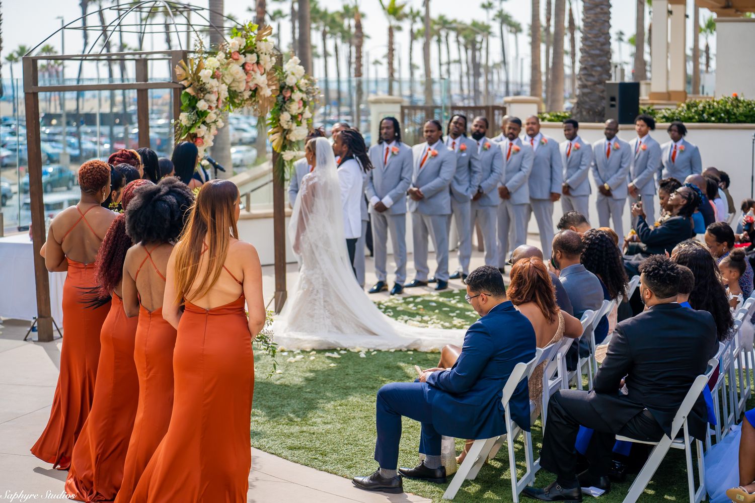 Bridesmaids in orange dresses walk toward bride and groom at outdoor waterfront wedding ceremony.