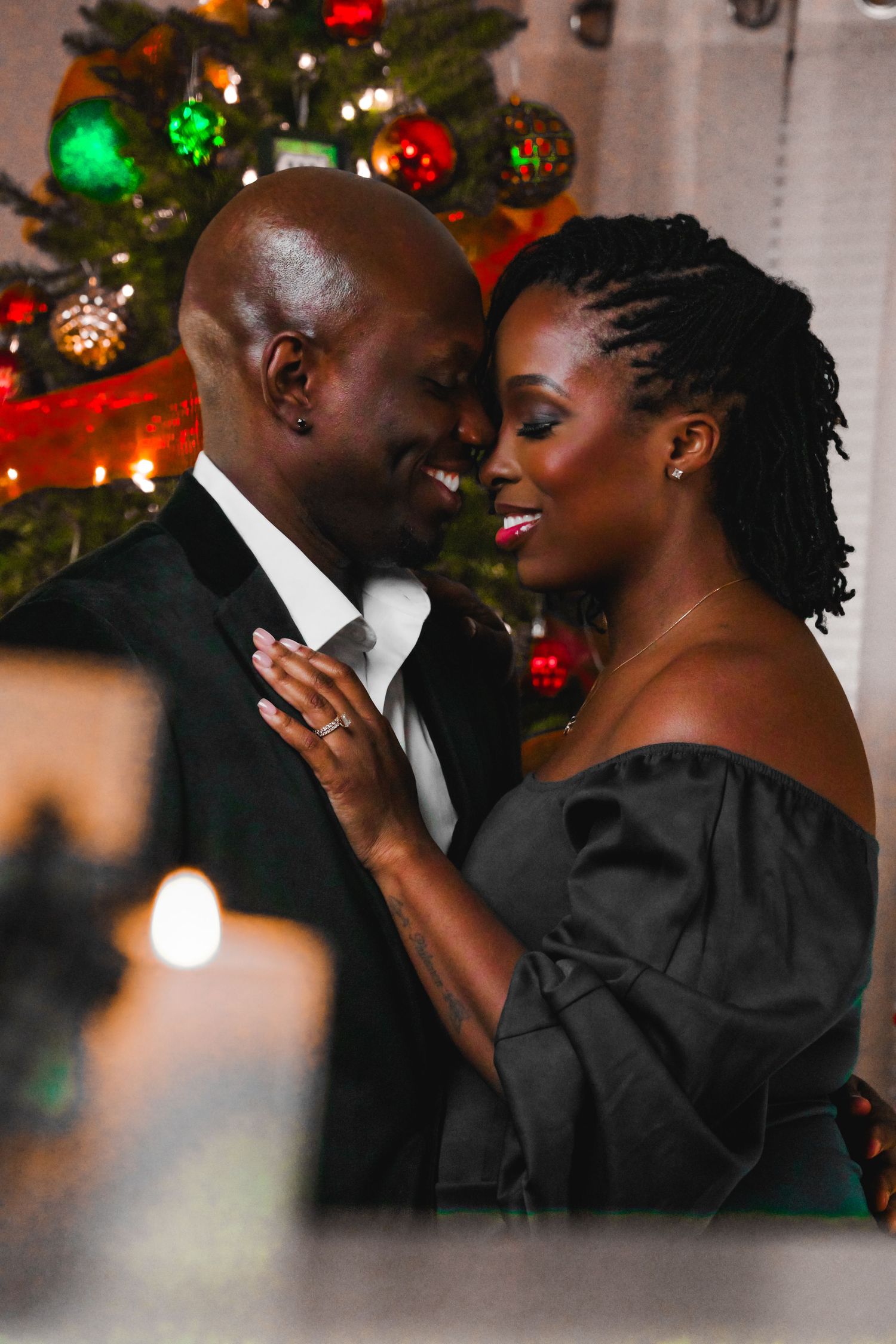 Elegant couple in black formal attire sharing a smile in front of a festive Christmas tree.