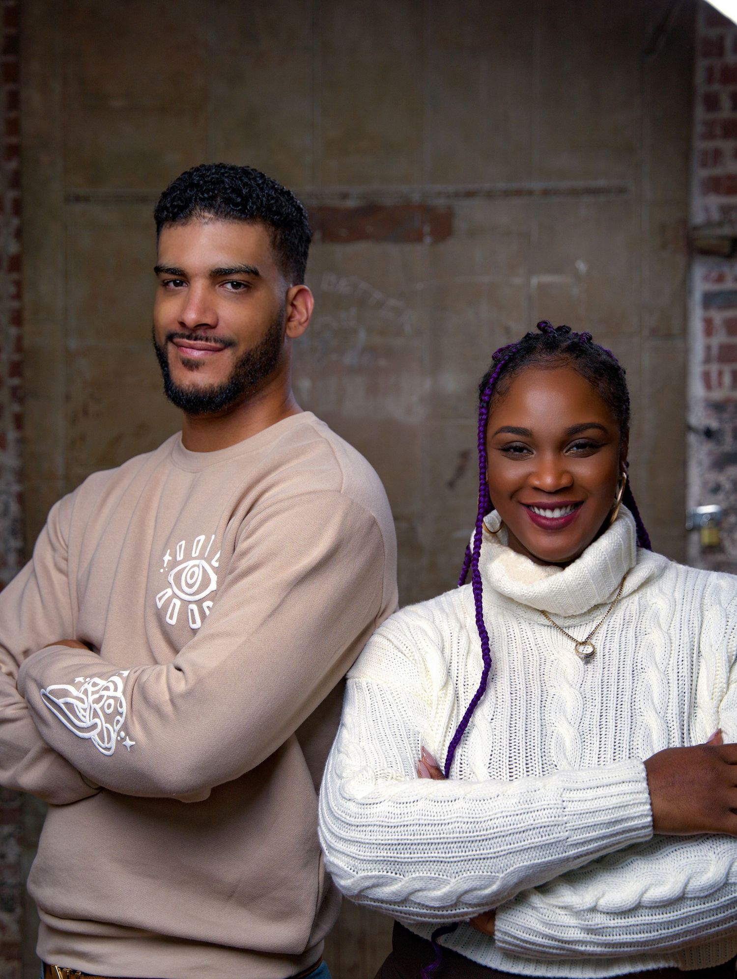 Confident couple standing with arms crossed, man in beige sweatshirt, woman in white turtleneck.
