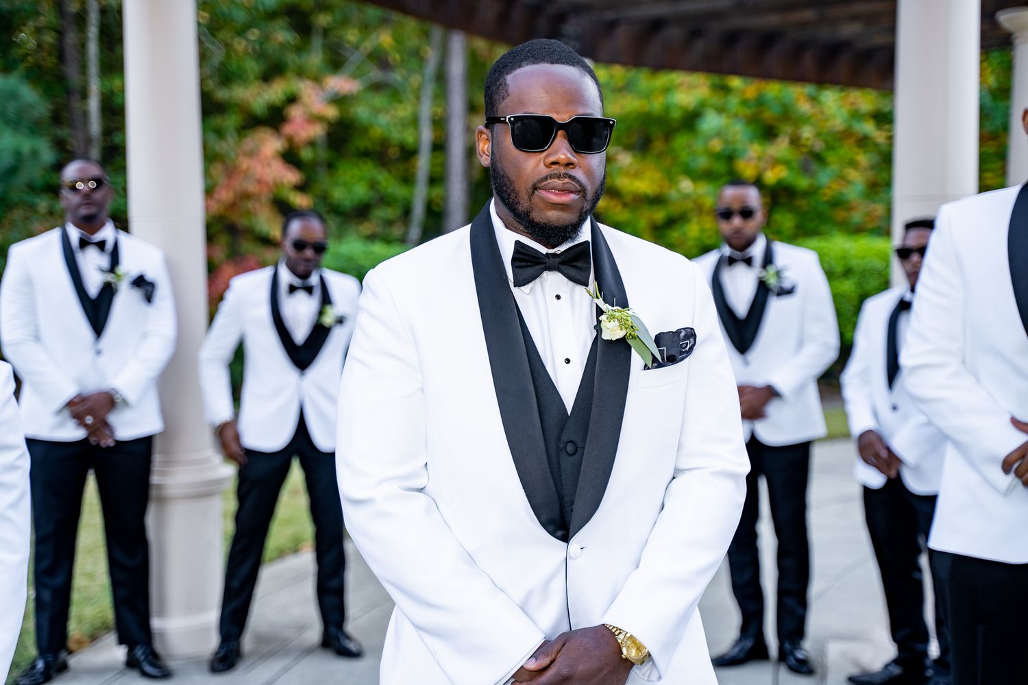 Groom in white tuxedo with black lapels posing confidently with groomsmen in matching suits outdoors.