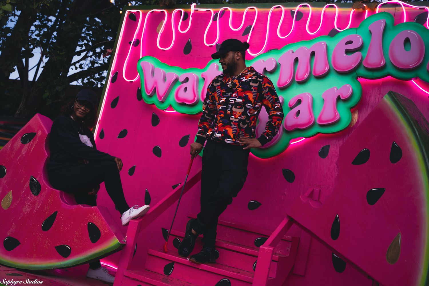 Person in colorful shirt posing at vibrant pink watermelon-themed outdoor installation at night.