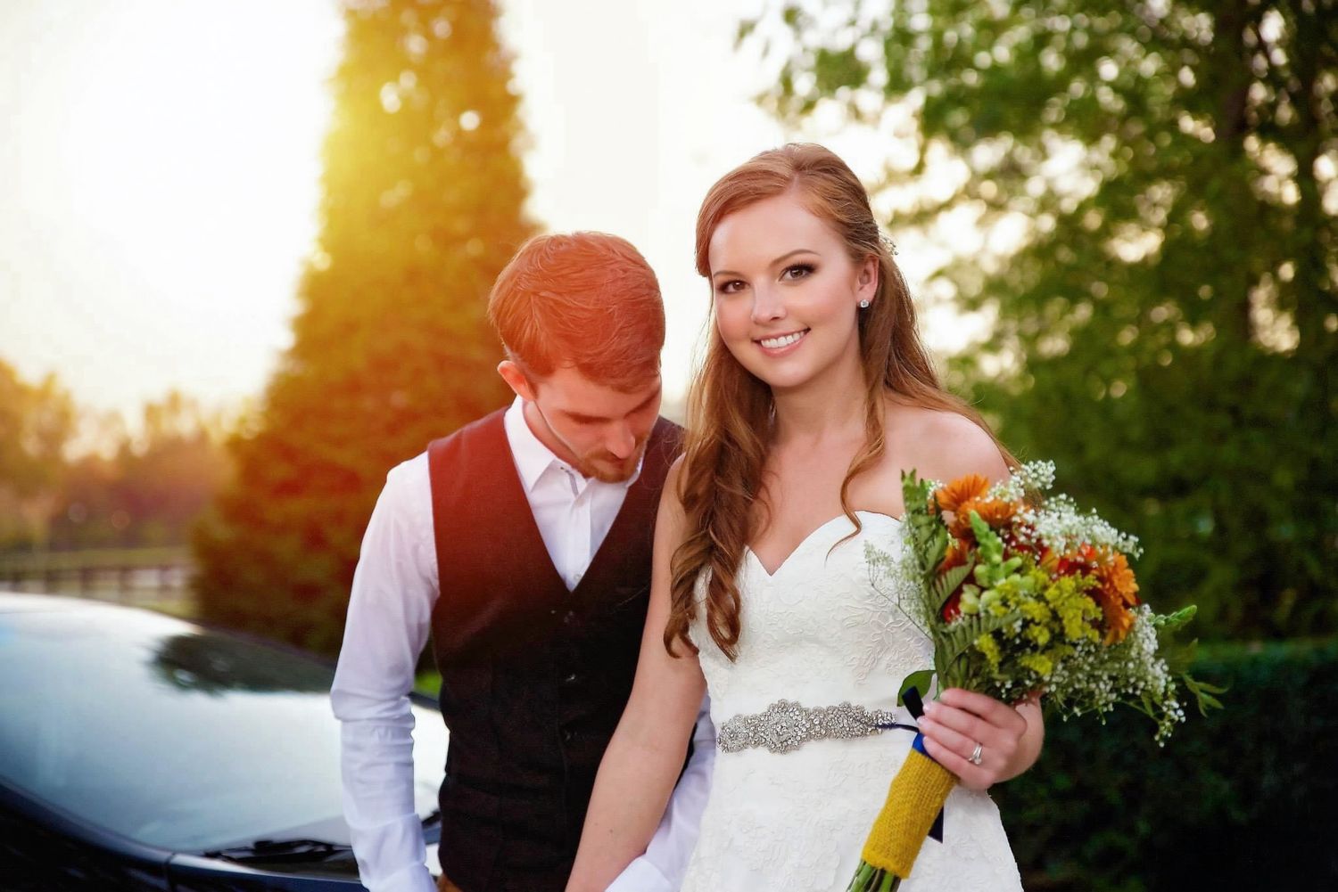 Bride holding colorful bouquet smiles beside groom in vest during outdoor wedding photo session at sunset.