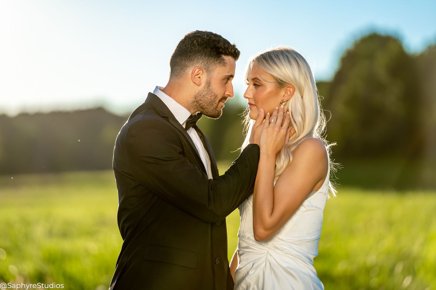 Groom tenderly holding brides face as they share a romantic moment in a sunny meadow.