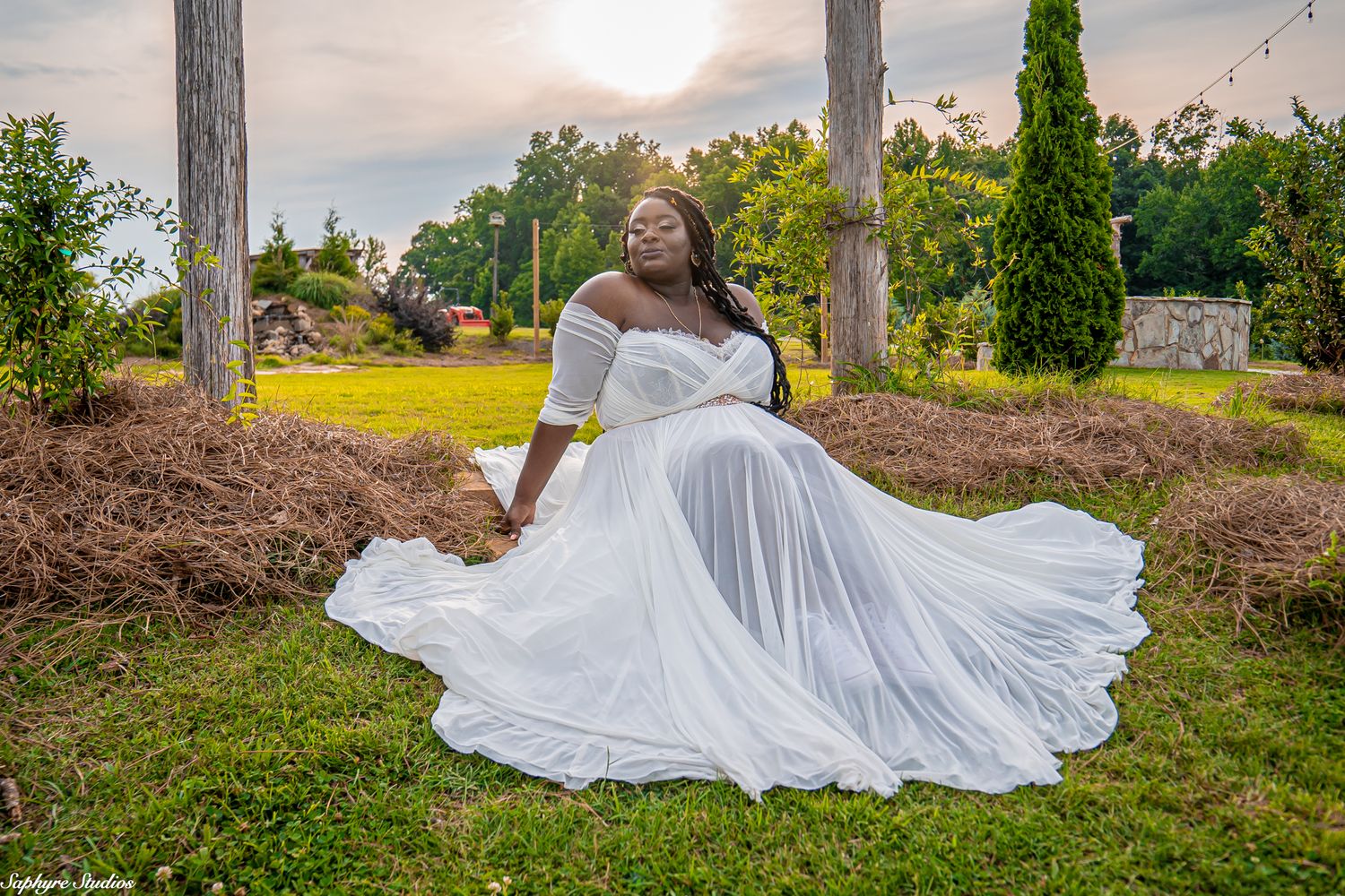 Bride in flowing white gown poses gracefully outdoors at sunset in a lush green garden setting.