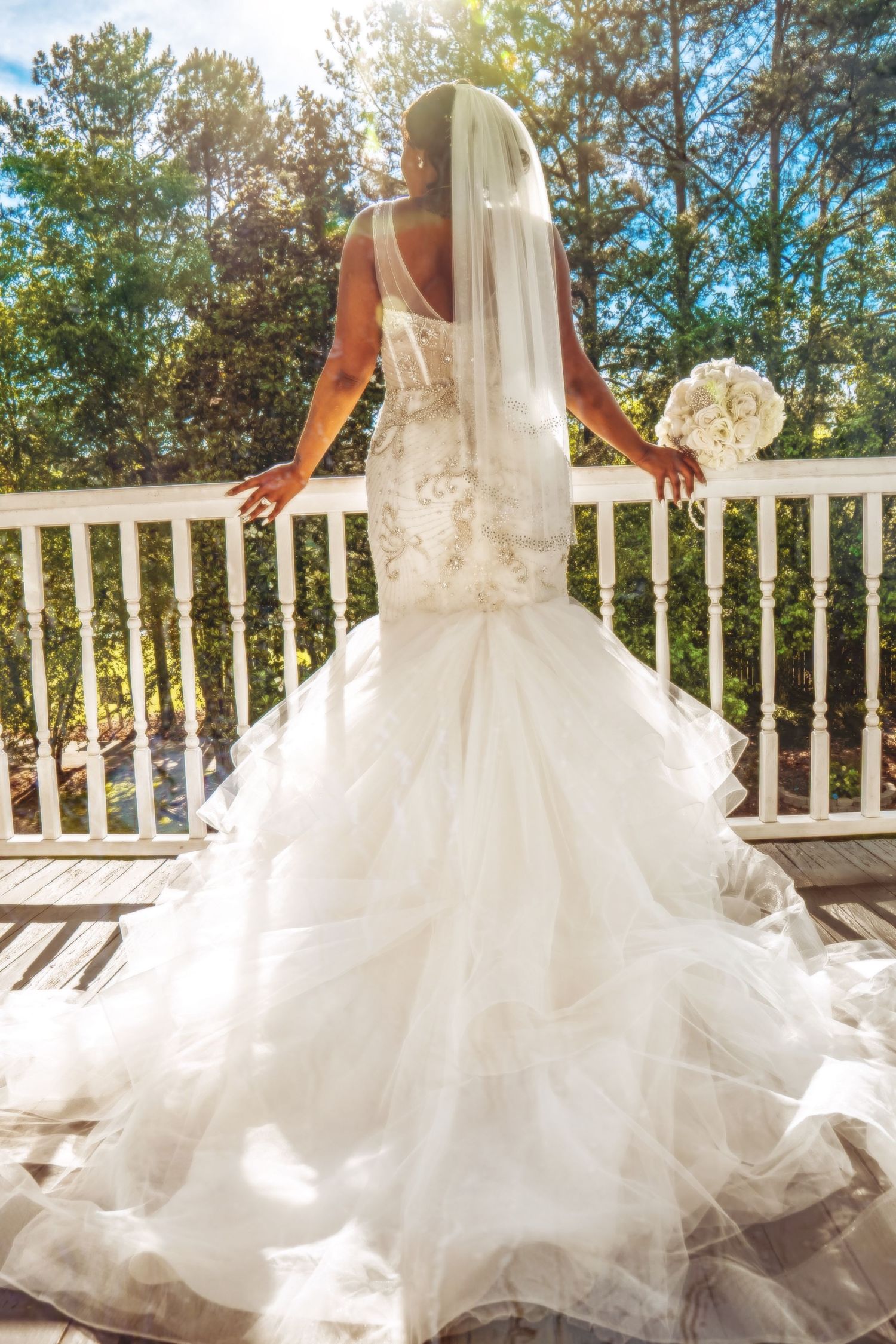Bride in mermaid gown with veil and bouquet poses on sunlit deck with trees behind her.
