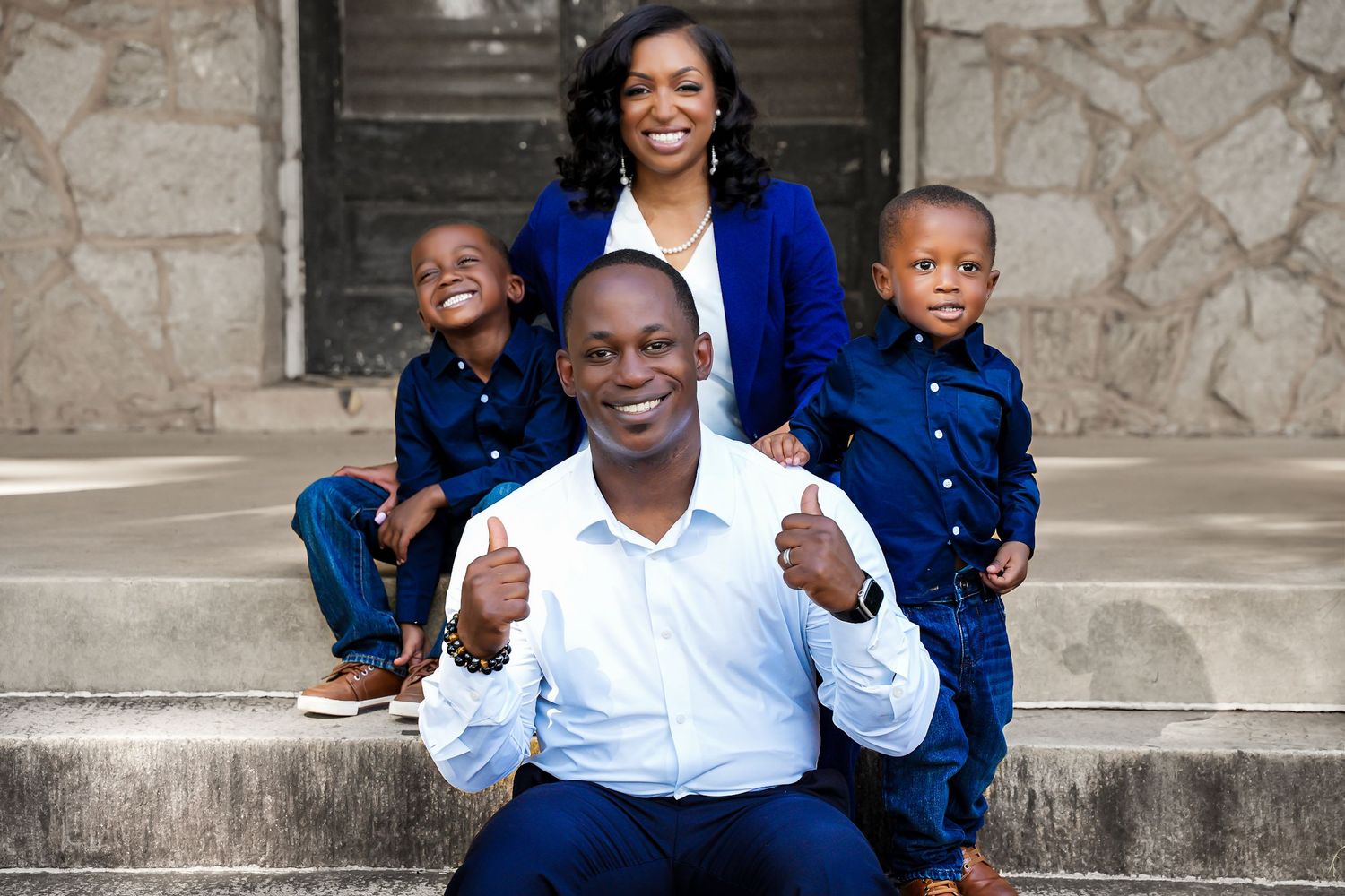 African American father giving thumbs up while family poses playfully on stone steps in blue outfits.