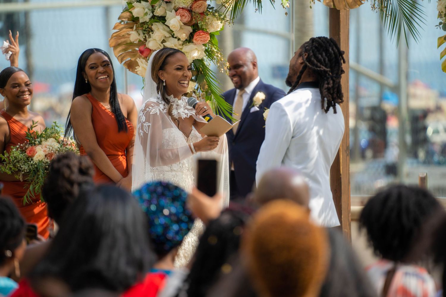 Bride in beaded gown speaks vows to dreadlocked groom under floral arch at outdoor wedding.