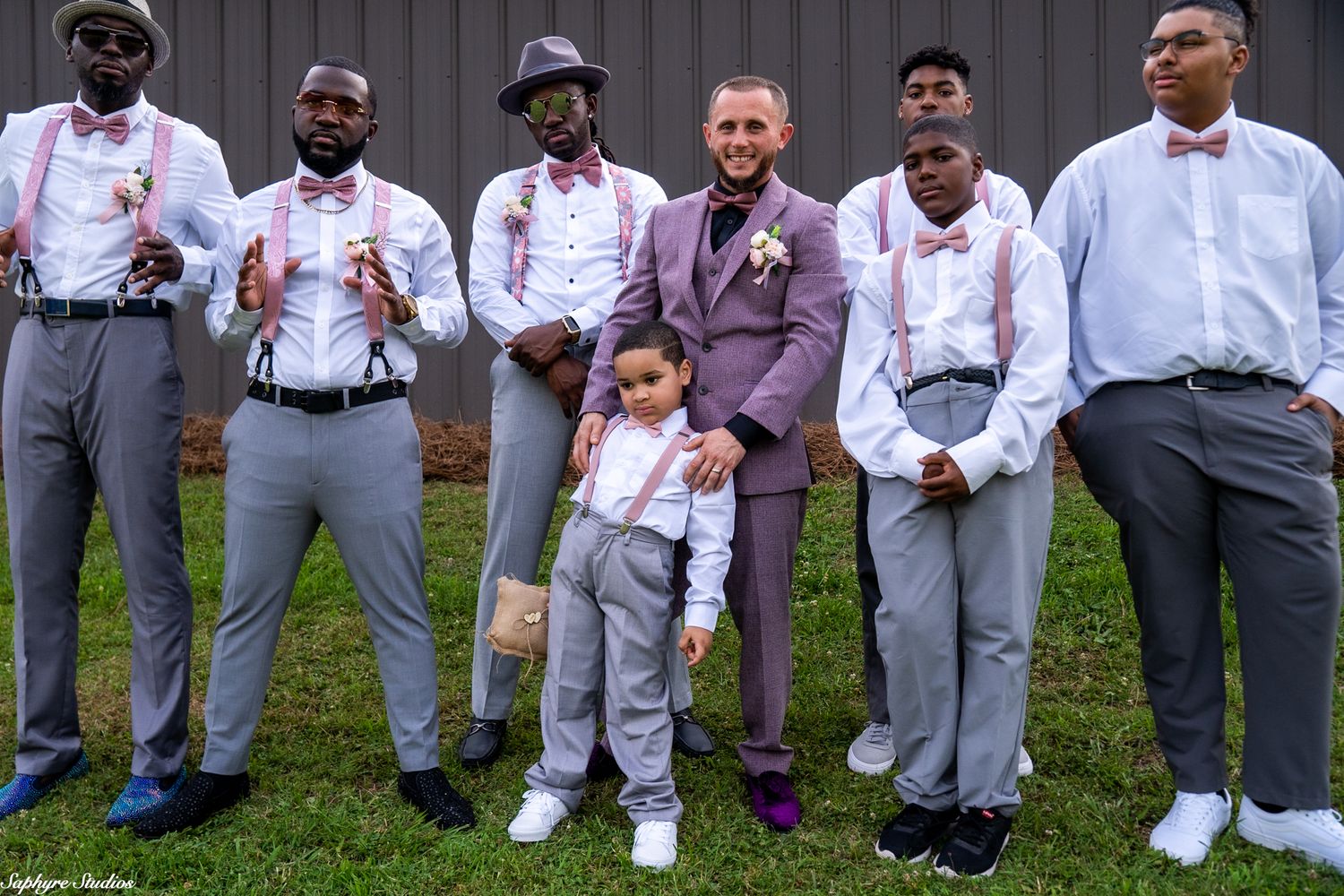 Groom in purple suit poses with groomsmen in gray suits and pink suspenders with young ring bearer on lawn.