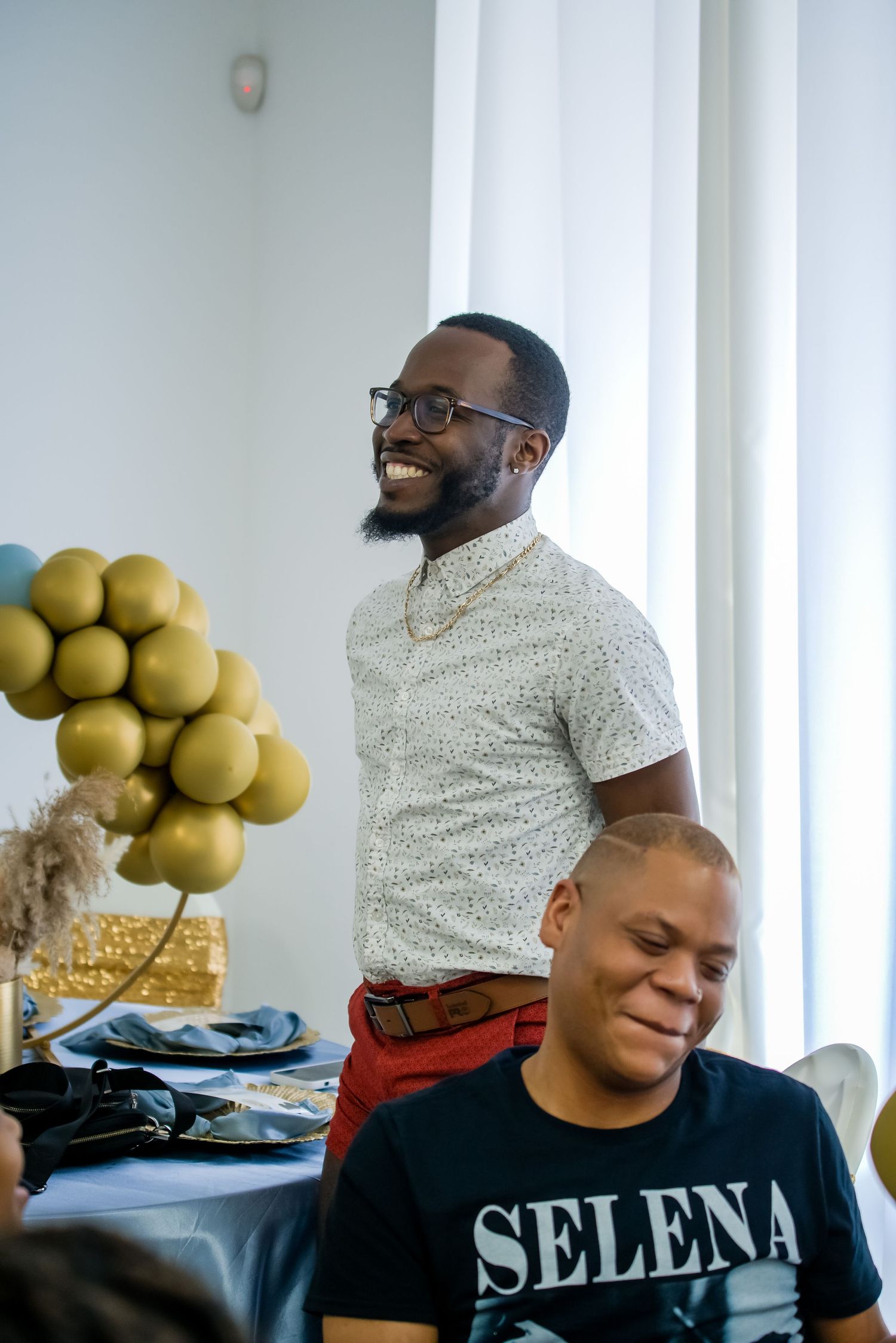 Smiling Black man in glasses at celebration event with gold balloons in background.