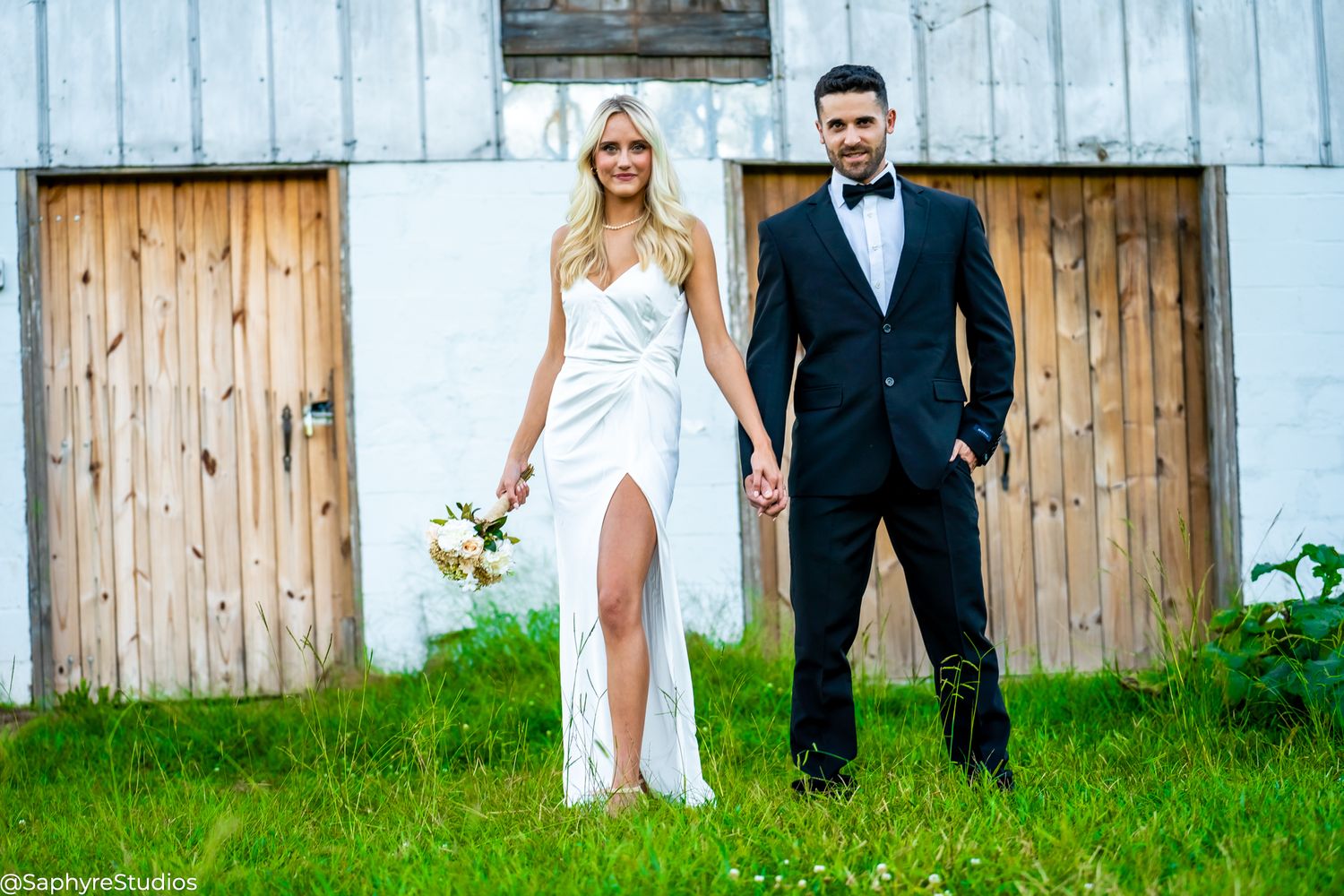 Bride and groom hold hands in front of rustic barn doors in wedding attire.