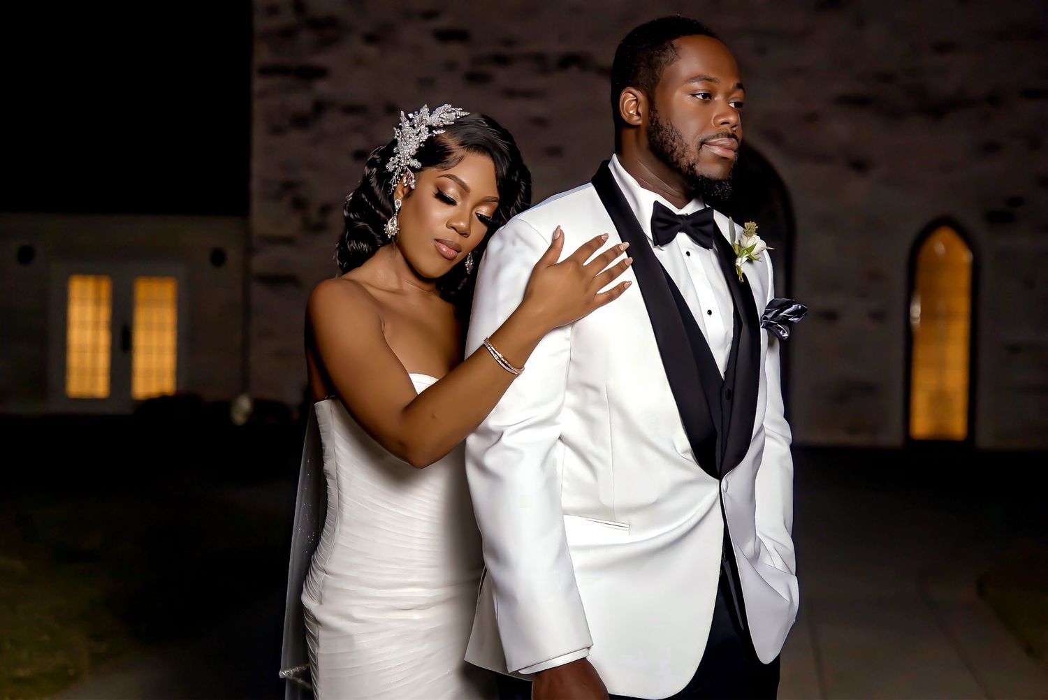 Bride in sleek gown leans on groom in white tuxedo jacket during dramatic nighttime wedding portrait.