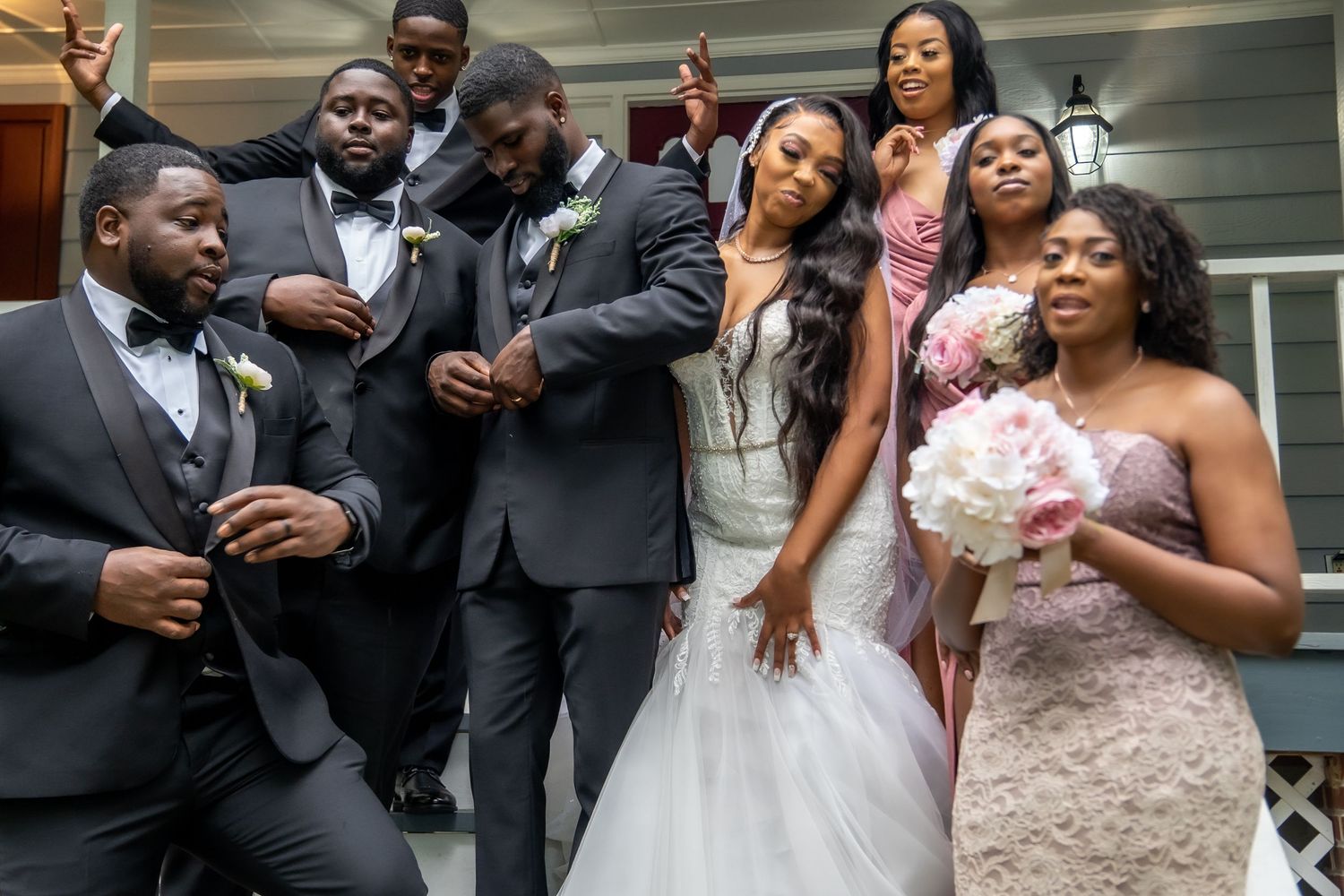 Lively wedding party celebration on porch with bride holding pink bouquet in white gown.