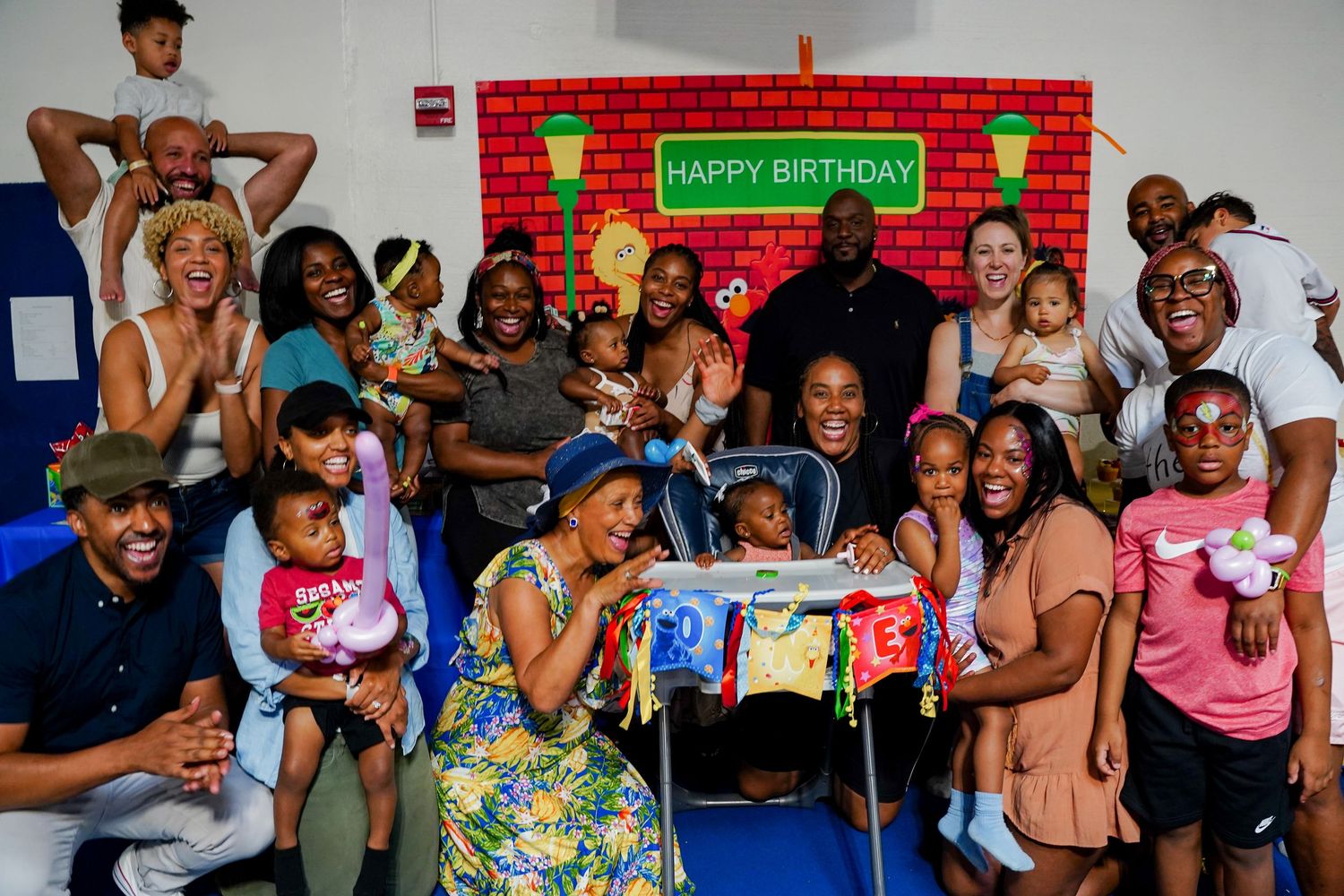 Large group celebrating child's birthday party in front of colorful Sesame Street themed backdrop.