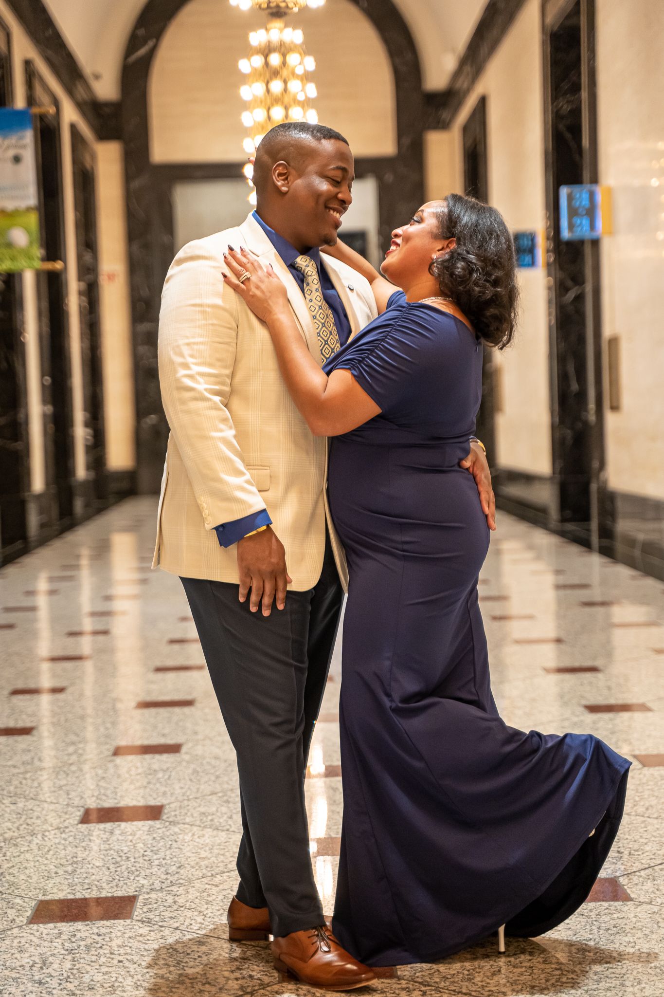 Smiling couple embraces in elegant hallway, man in cream blazer, woman in navy blue formal dress.