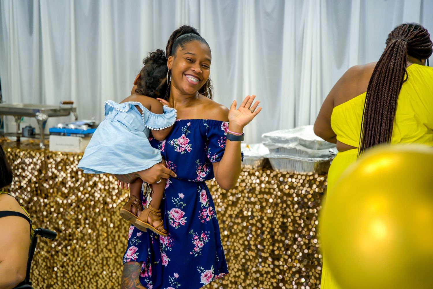 Joyful woman in floral dress holds young child at a gold sequin backdrop party.