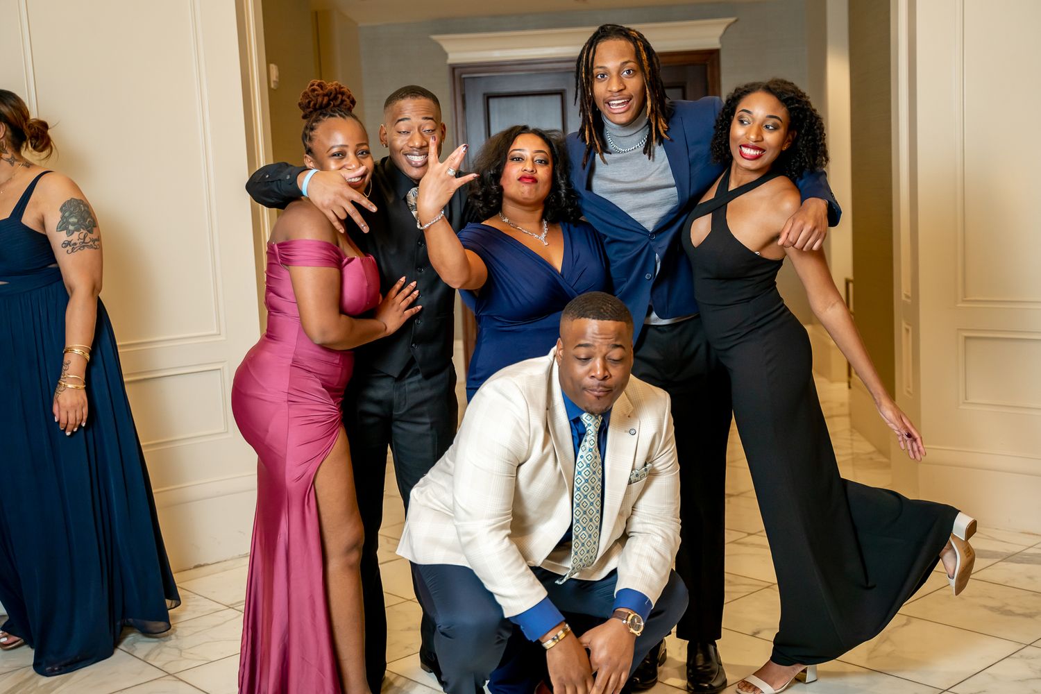 Laughing group in elegant formal attire posing playfully in a well-lit hallway at a gala.