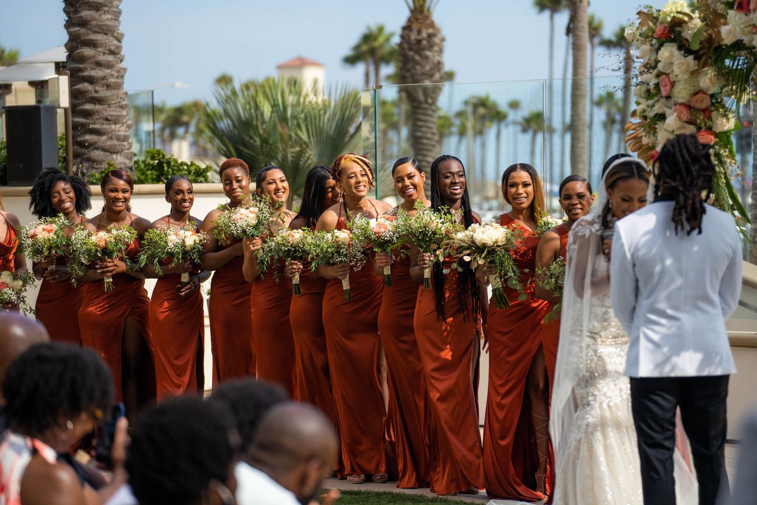 Bridesmaids in rust orange gowns holding bouquets line up at an outdoor beach wedding ceremony.