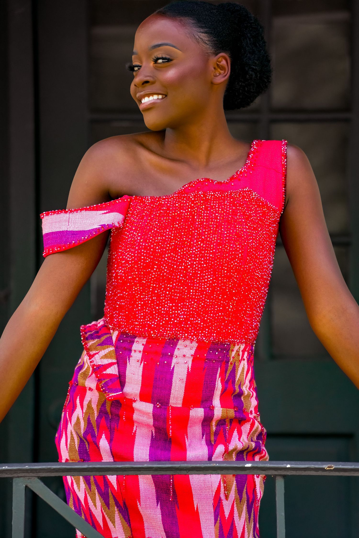 A beautiful woman in a red beaded and colorful African print dress smiles outdoors.