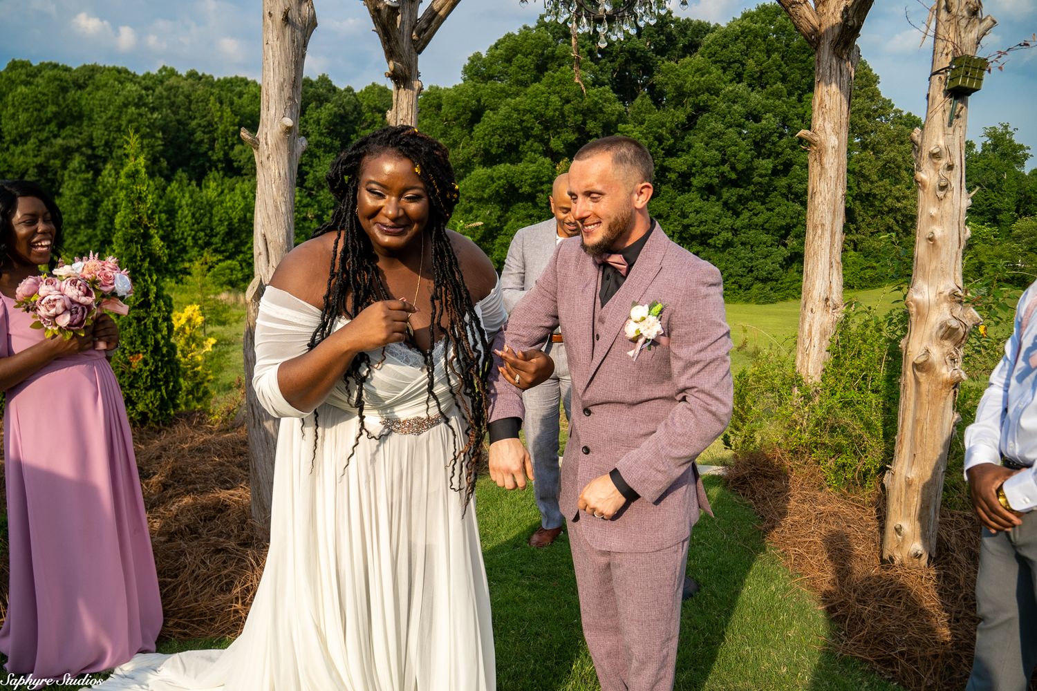 Joyful newlywed couple laughs and holds hands after outdoor wedding ceremony with bridesmaids in pink nearby.