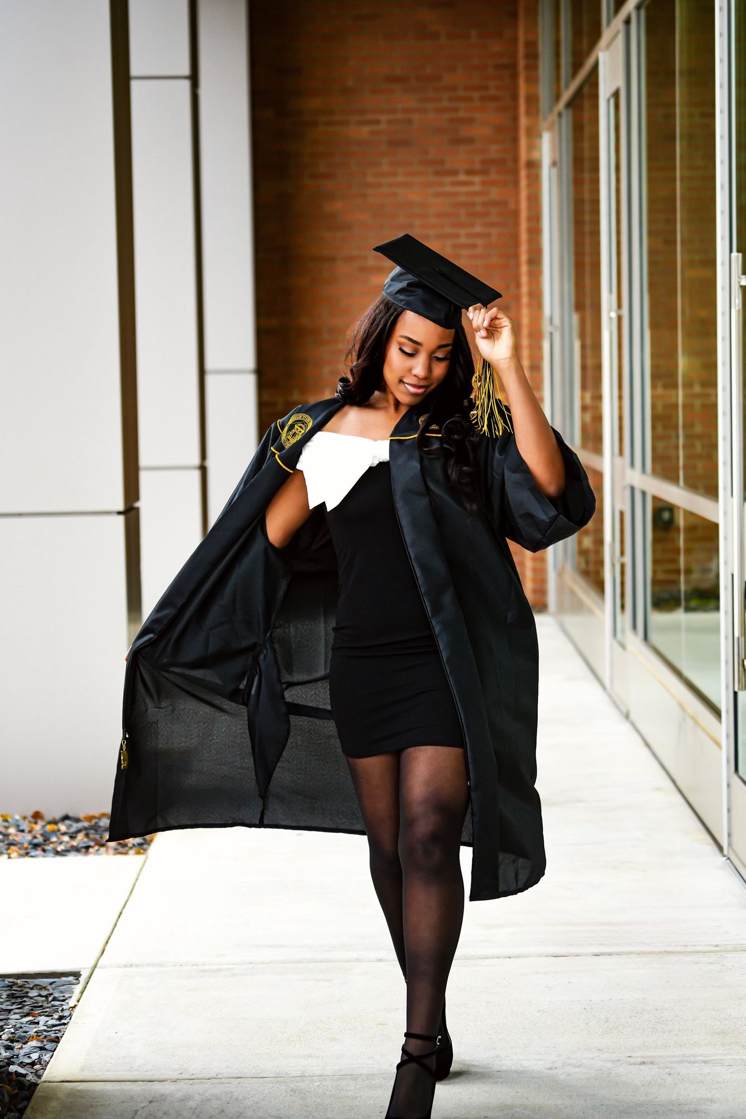 Young Black woman in graduation cap and gown walking outdoors near brick building.