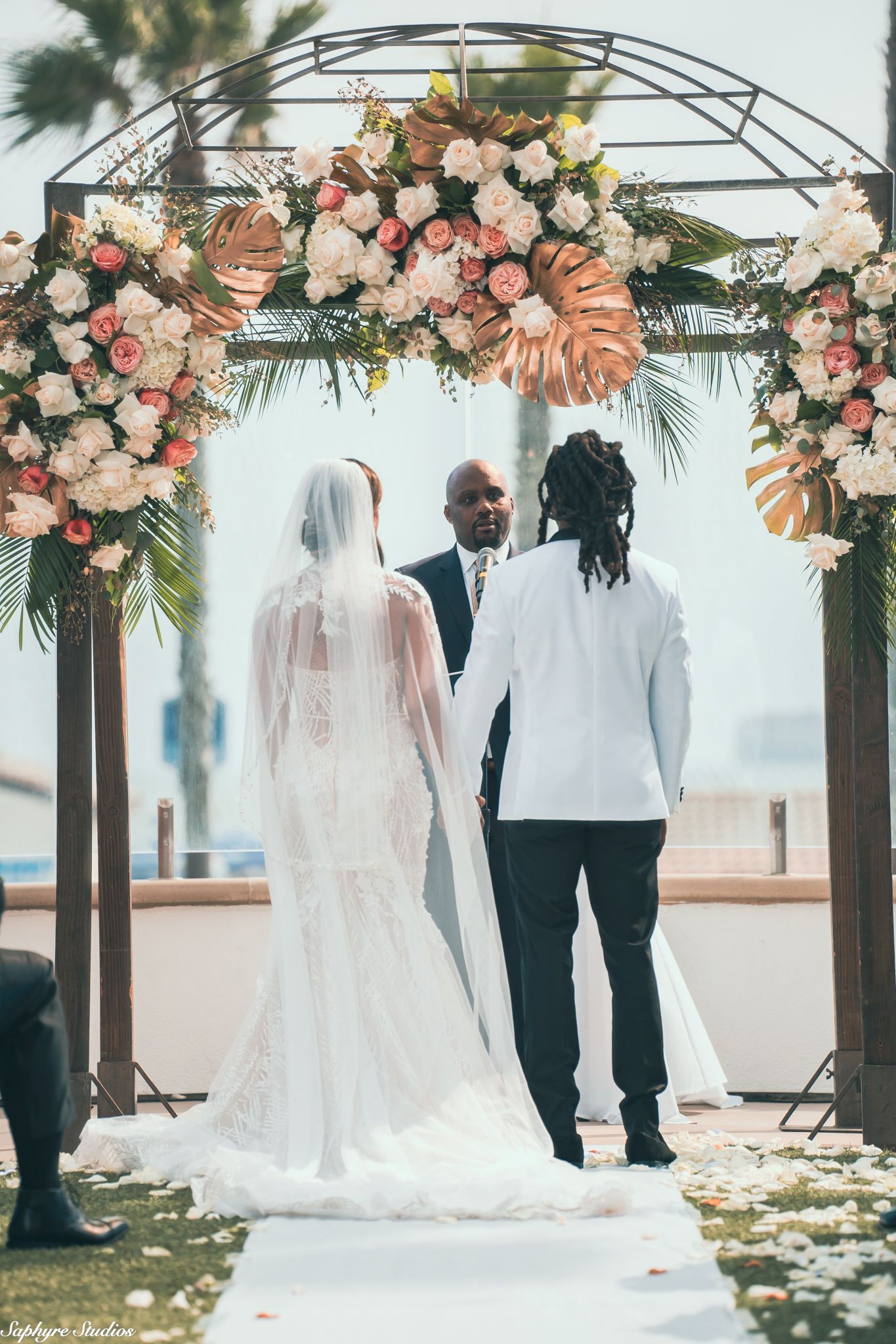 Bride in lace gown and veil stands with groom under floral arch at outdoor wedding ceremony.