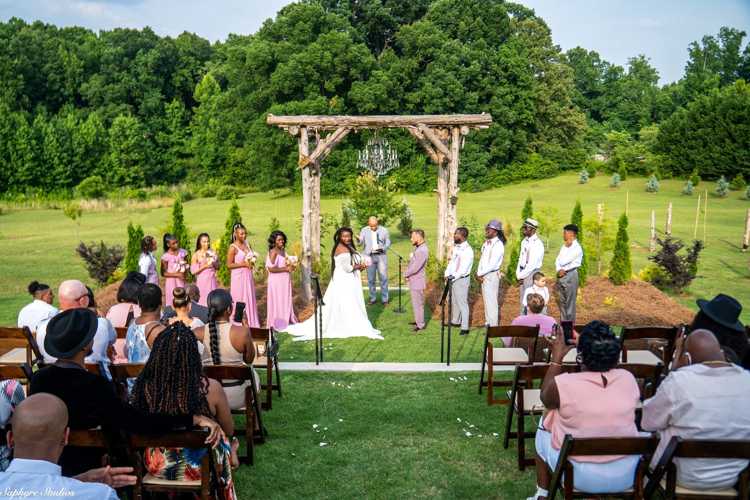 Outdoor wedding ceremony on lush green lawn with guests seated in wooden chairs facing rustic log arch altar.