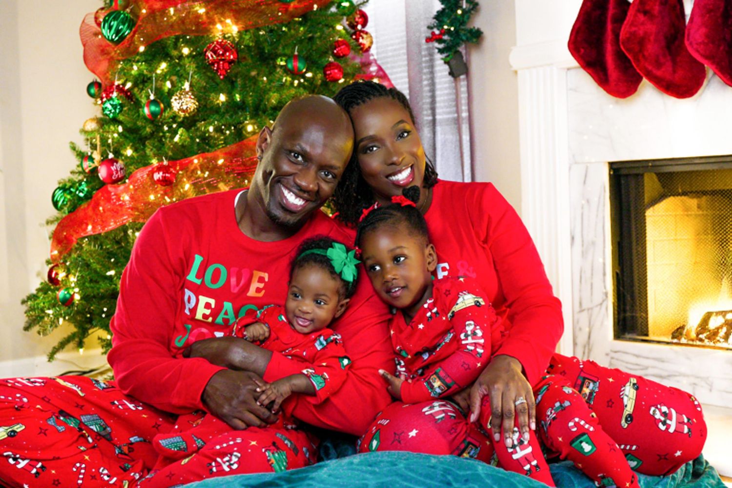 Family of four in matching red Christmas pajamas smiling by festive holiday tree.