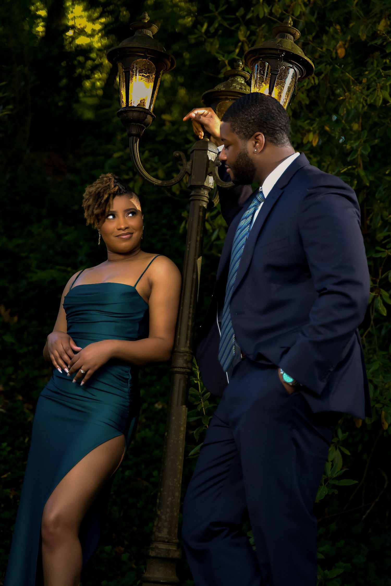 Couple posing by vintage lamp post, woman in teal gown and man in navy suit, surrounded by lush greenery.