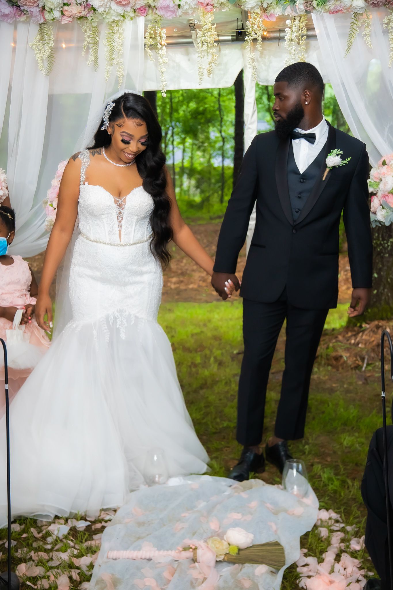 Bride in mermaid gown and groom in black tuxedo hold hands at outdoor wedding ceremony.