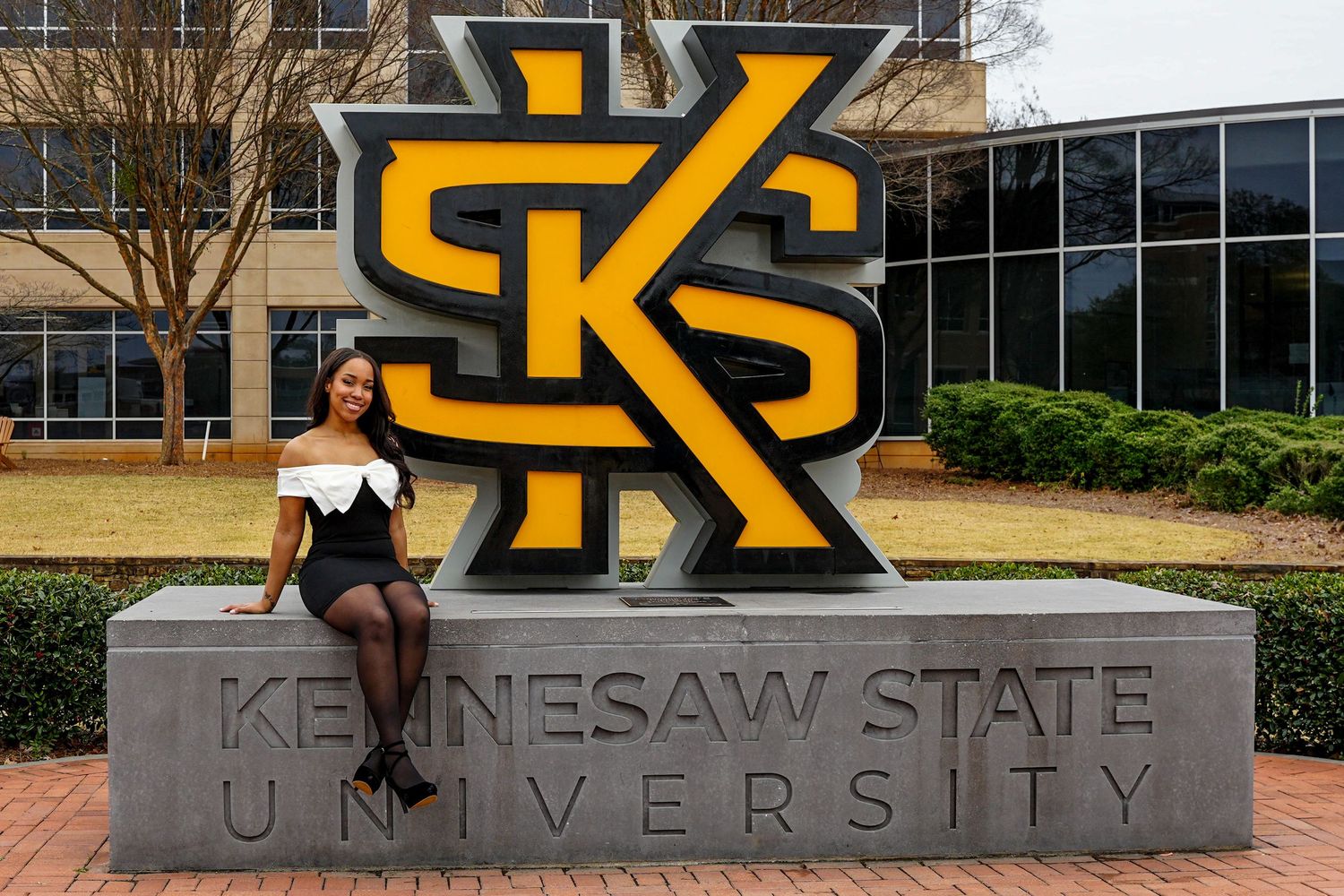Nursing graduate sits proudly in front of Kennesaw State University iconic black and gold campus sign.