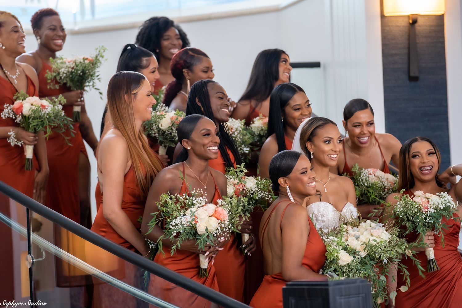 Large group of bridesmaids in rust orange dresses laughing and holding white floral bouquets on staircase.