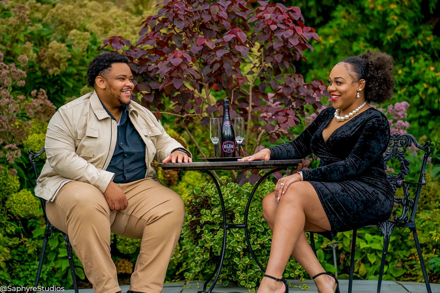 Couple sharing a romantic moment at outdoor garden table with wine bottle, dressed in elegant casual attire.