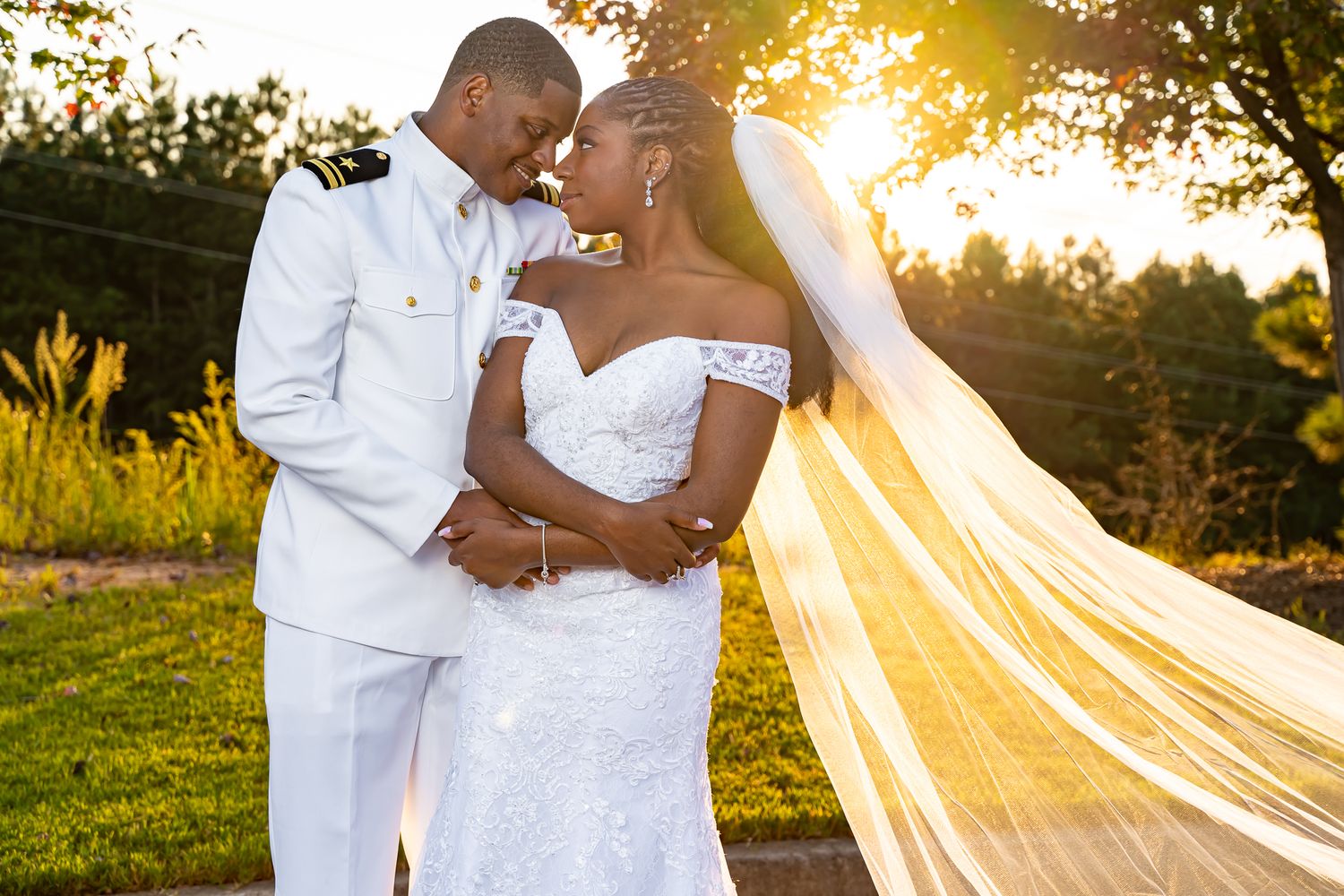 Military groom in white dress uniform embracing bride in lace gown during golden sunset outdoor wedding.