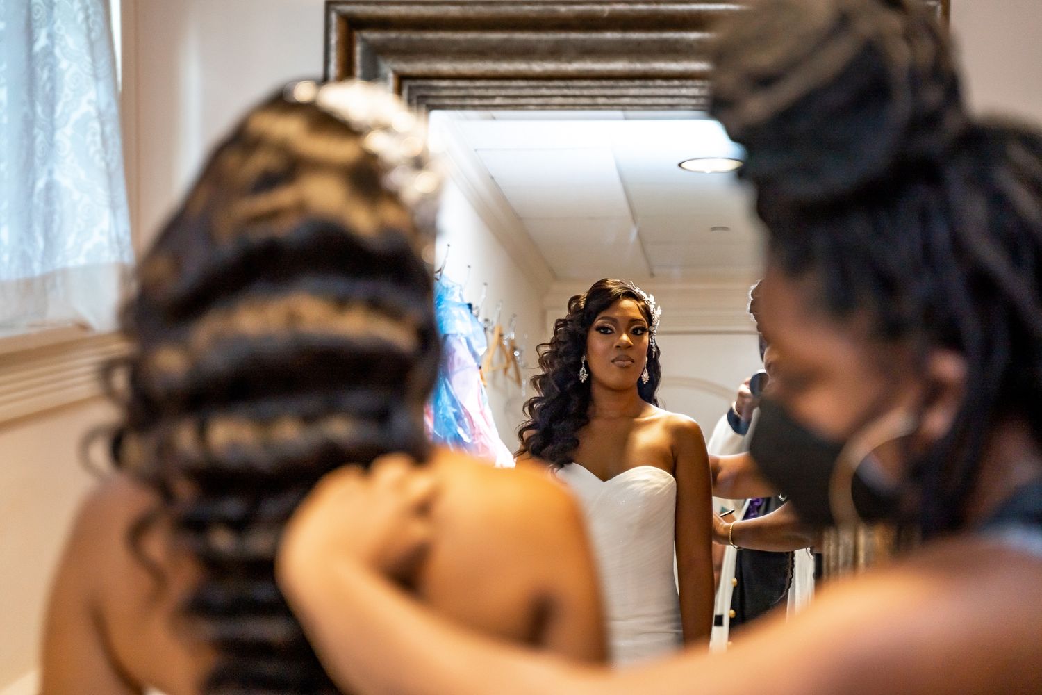 Bride in white gown gets hair styled by assistants while viewing reflection in mirror.