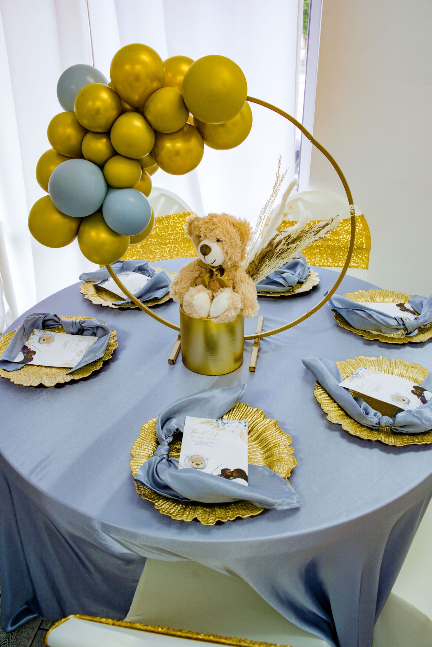 Elegant baby shower table with gold balloon arch, teddy bear centerpiece, and blue place settings.