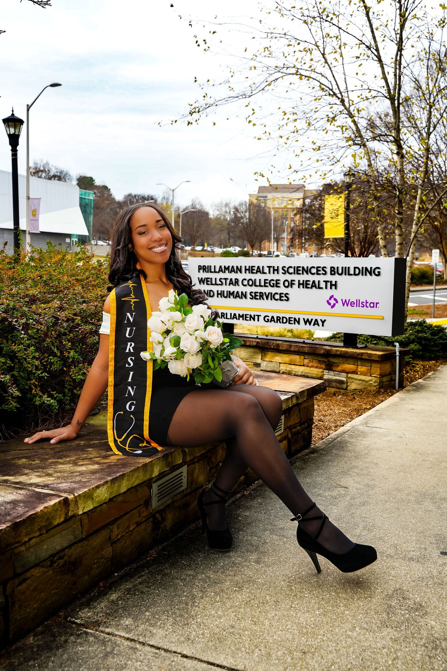Nursing graduate wearing black and gold sash holding white roses outside Wellstar College of Health building.