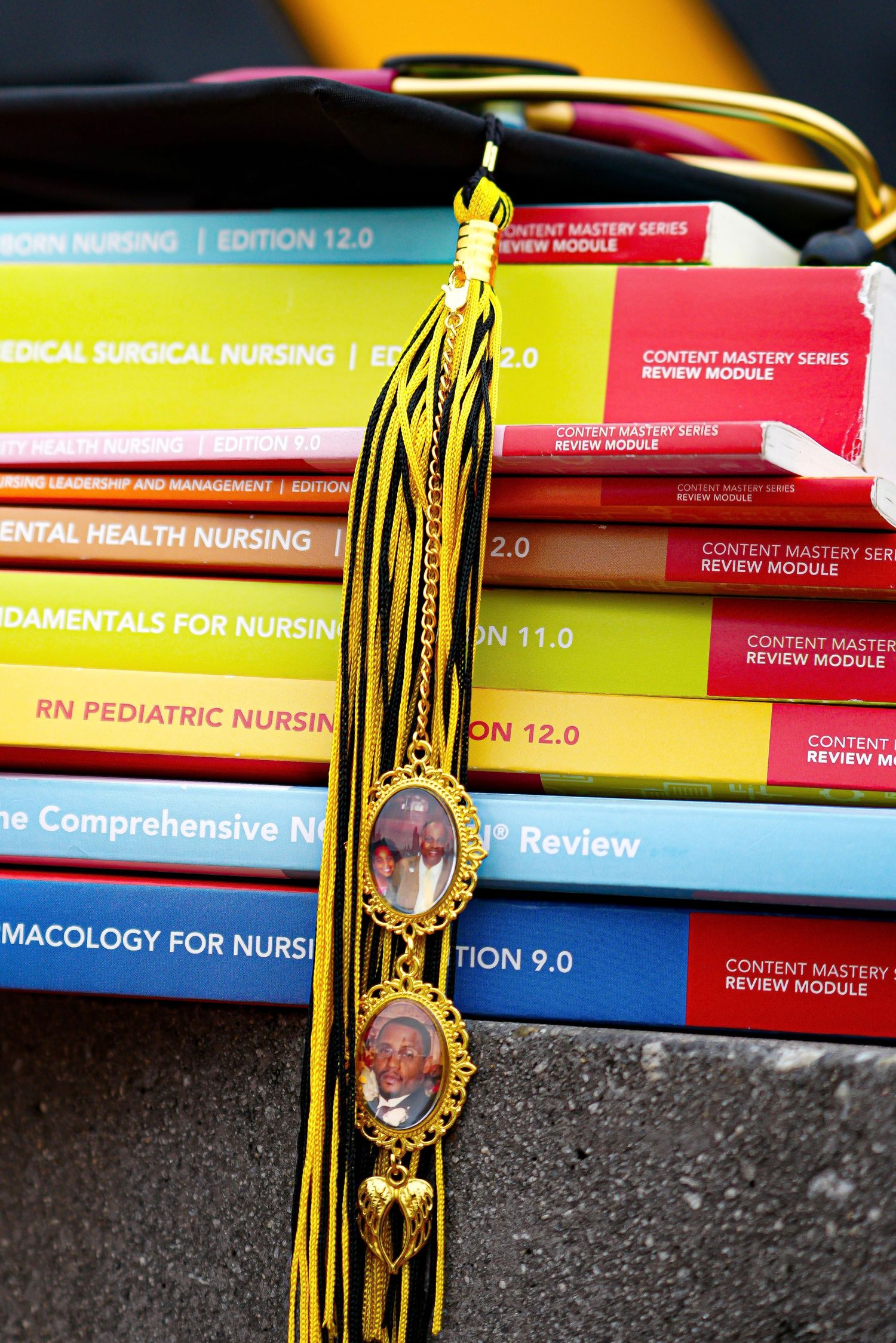Close-up of nursing graduation cap tassel with memorial lockets resting on colorful ATI nursing textbooks.