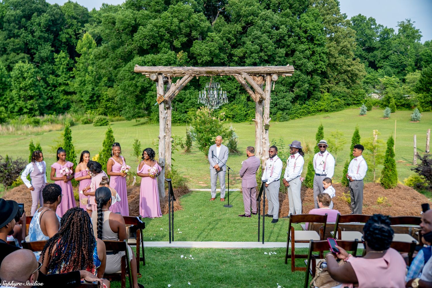 Wedding ceremony at rustic outdoor venue with bridesmaids in pink and groomsmen at the altar.