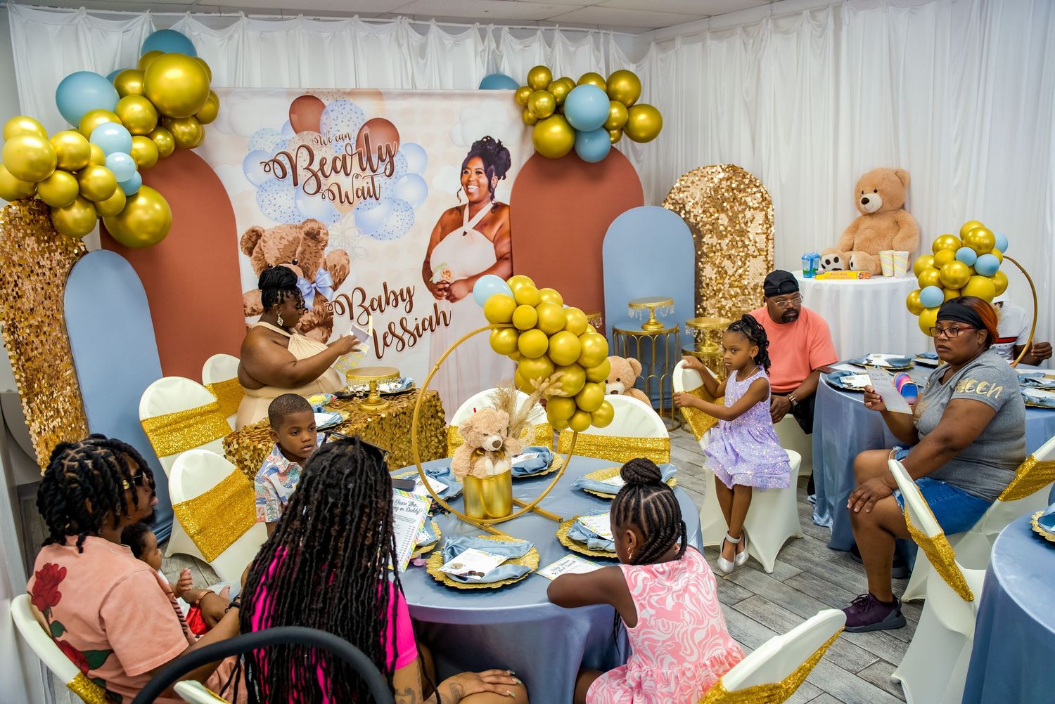 Guests celebrate at a gold and blue baby shower with balloon arch decor and large teddy bear centerpiece.