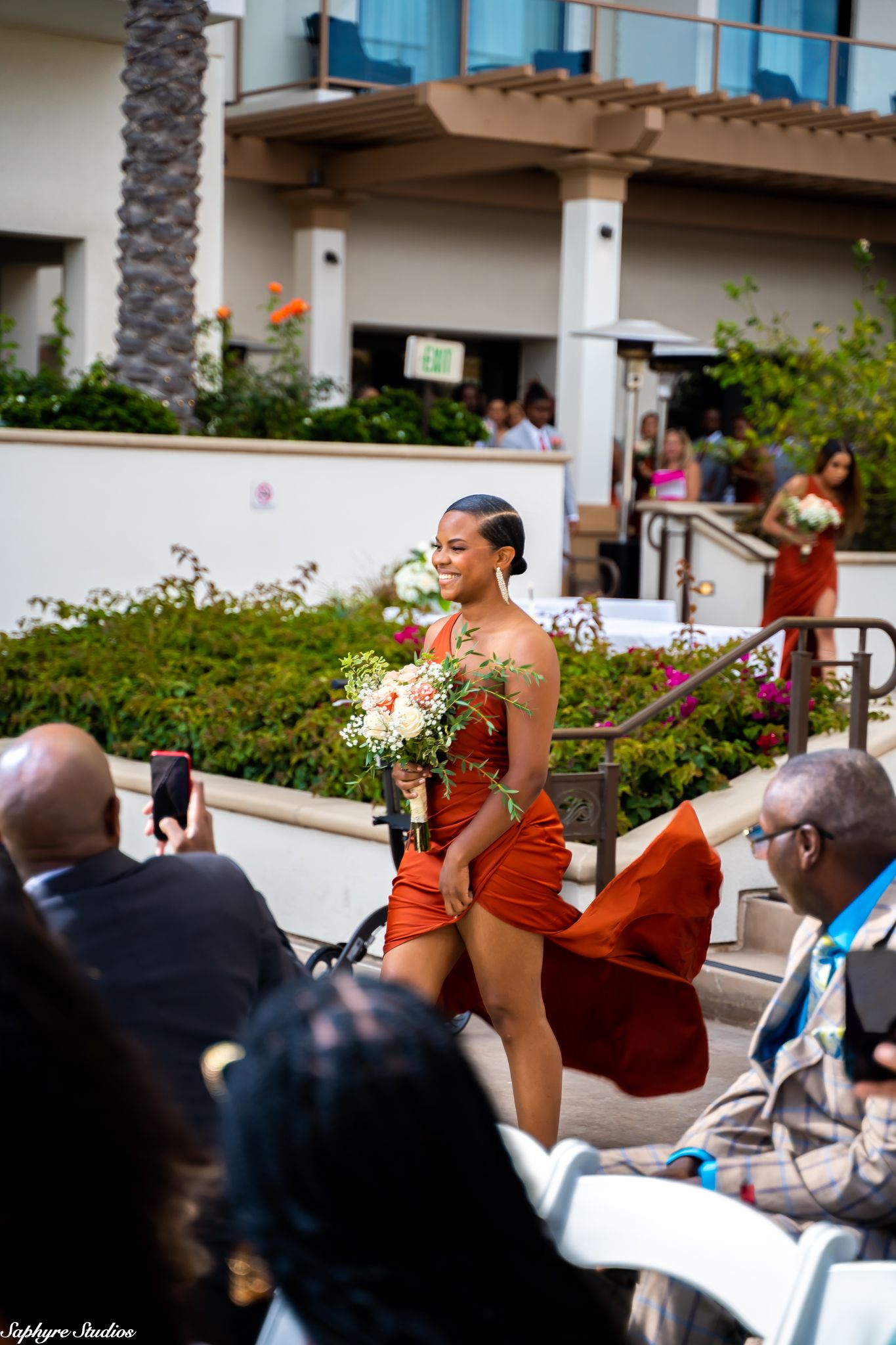 Bridesmaid in flowing rust orange dress carries bouquet while walking down outdoor wedding aisle.