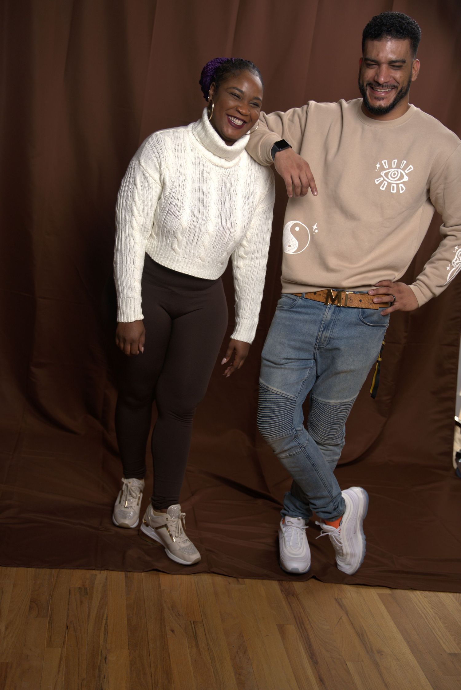Smiling couple posing against brown backdrop, woman in white sweater, man in beige sweatshirt.