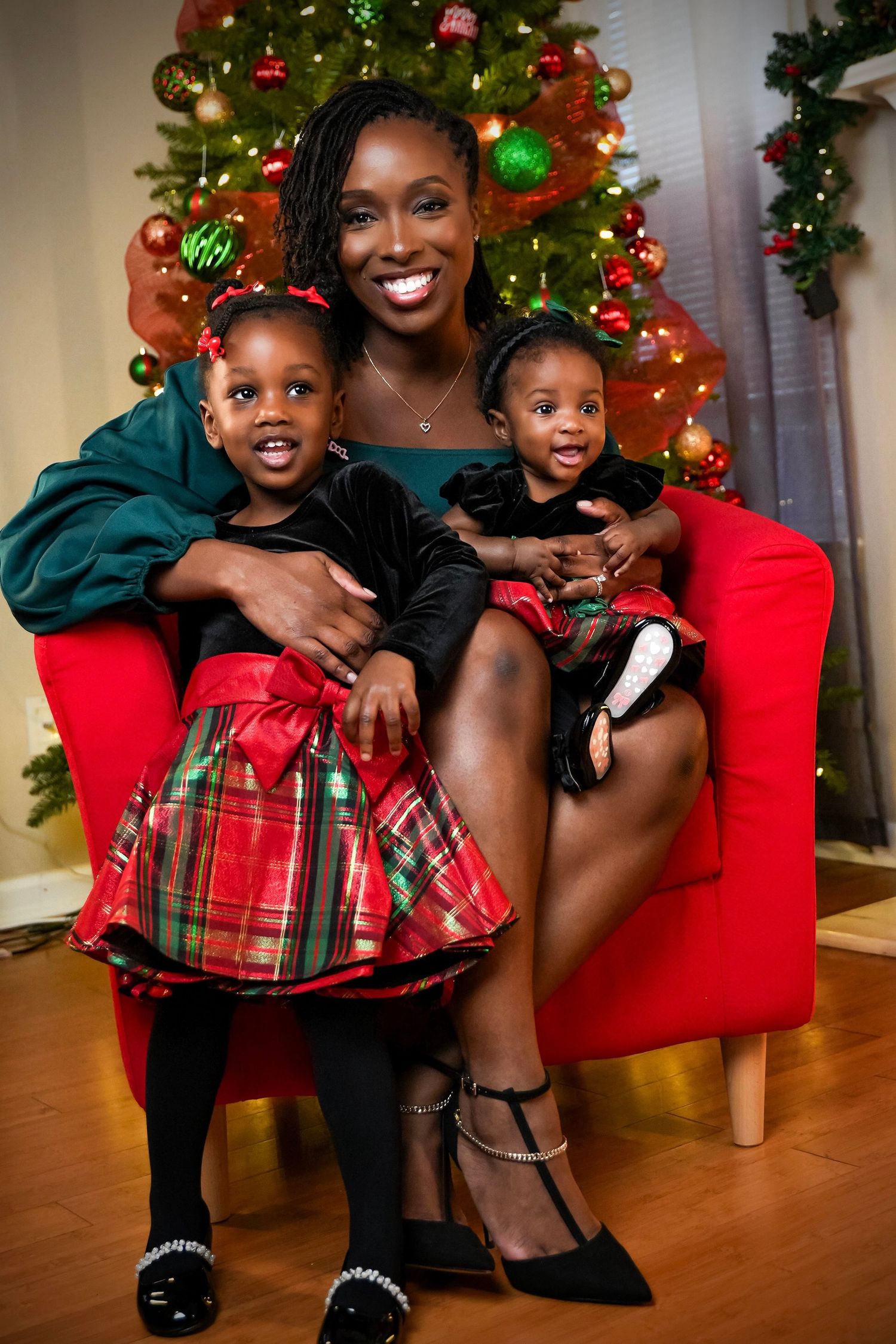 Mother smiling with two young daughters in holiday dresses seated in red chair.