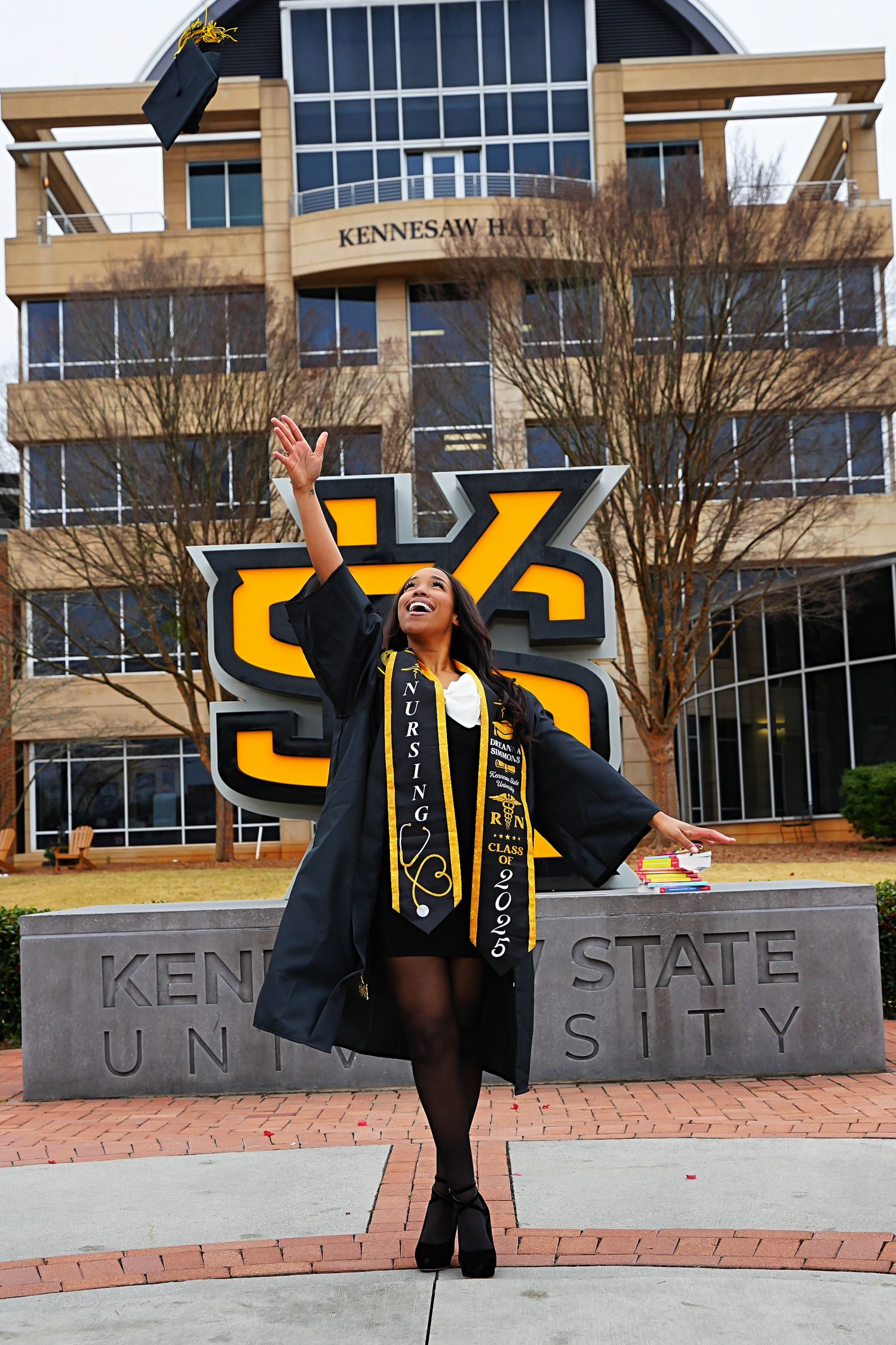 Excited graduate in cap and gown tosses cap in front of Kennesaw State University sign.
