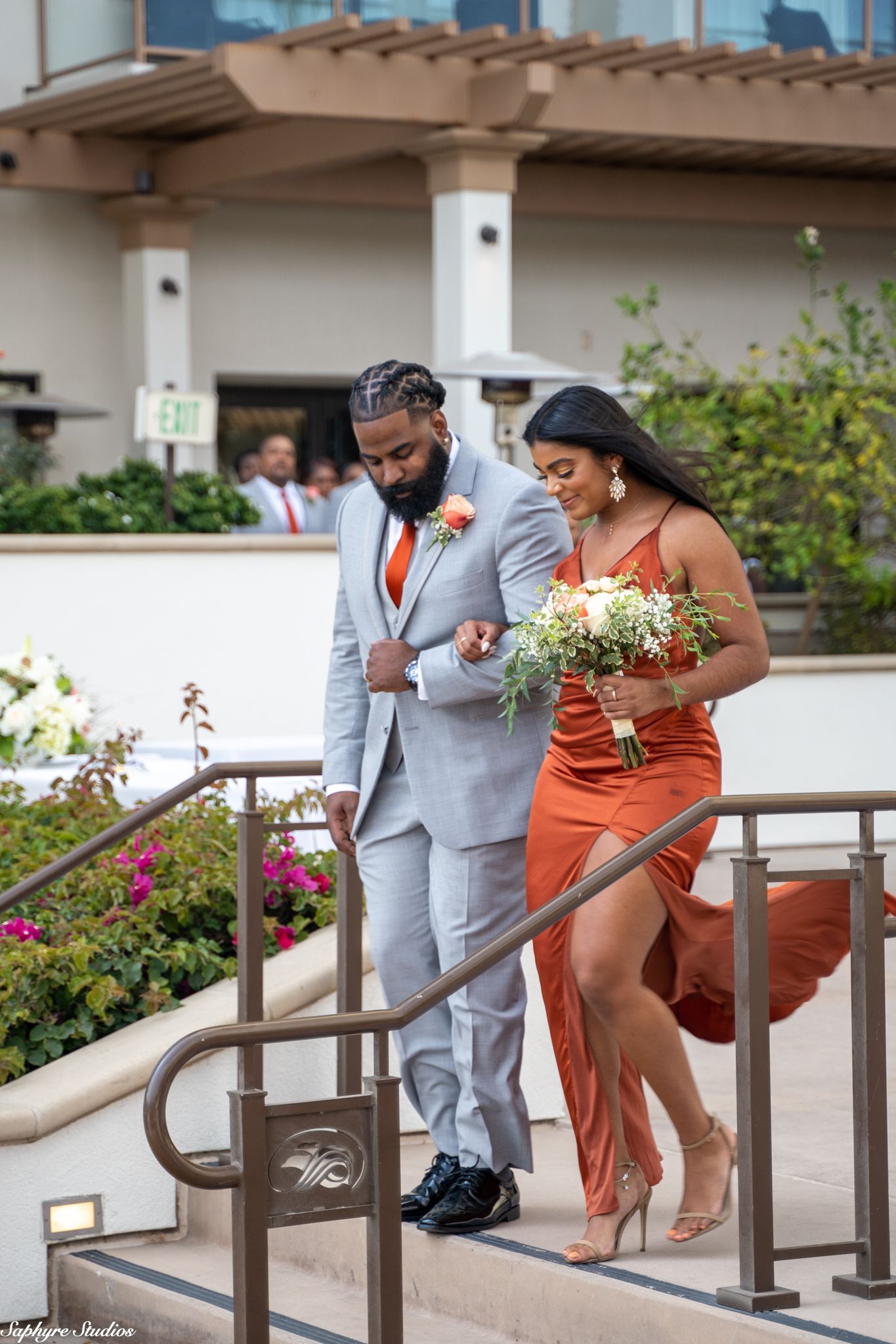 Groomsman in gray suit with orange tie escorts bridesmaid in rust dress down stairs at outdoor wedding.