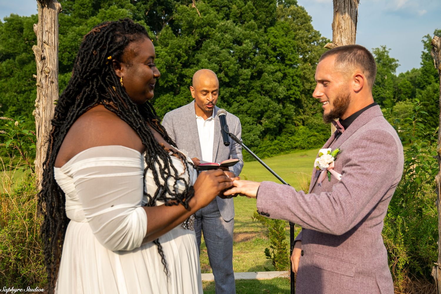 Groom places ring on brides finger during intimate outdoor wedding ceremony with officiant standing behind them.