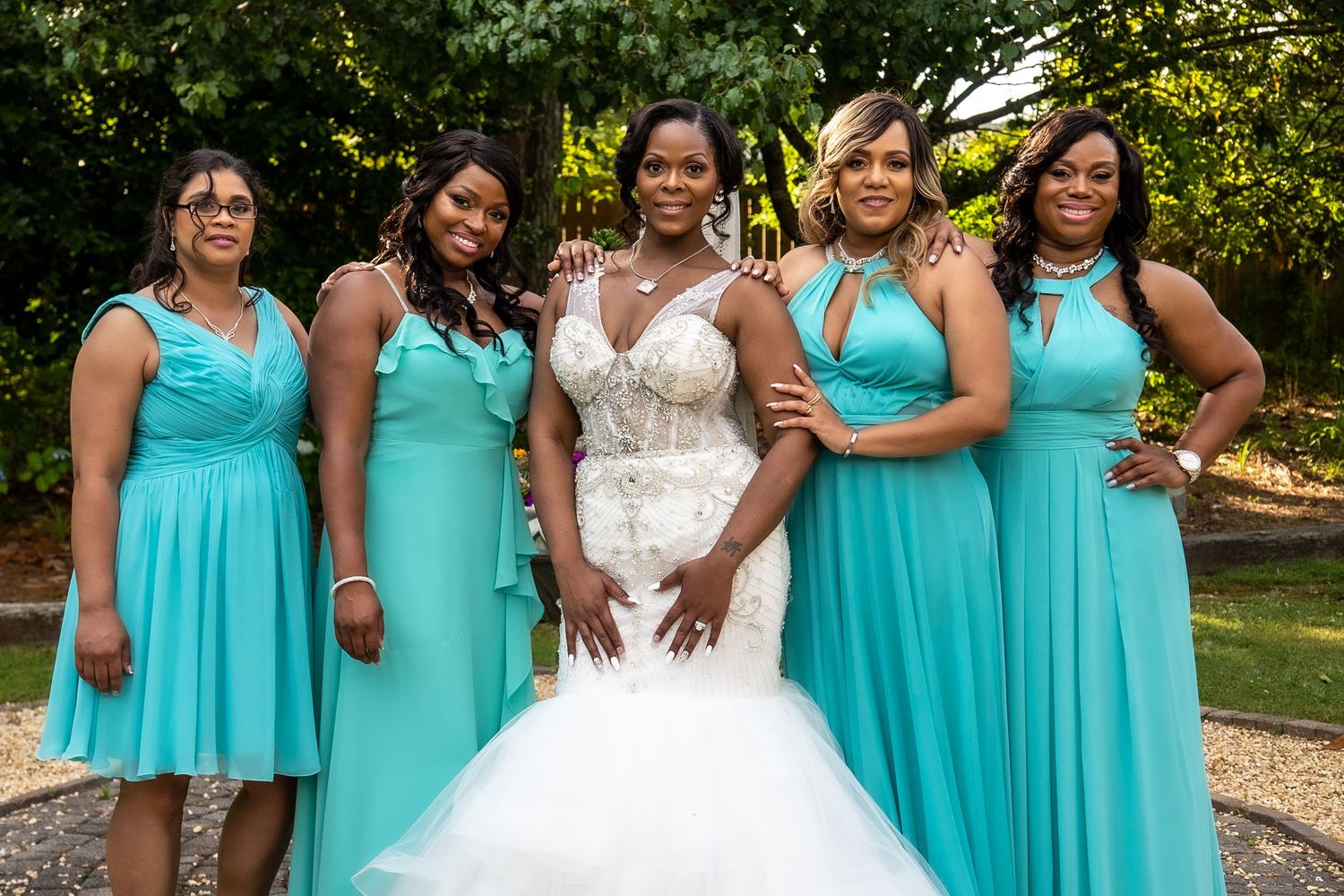 Bride in beaded mermaid gown stands with four bridesmaids in matching teal dresses outside.