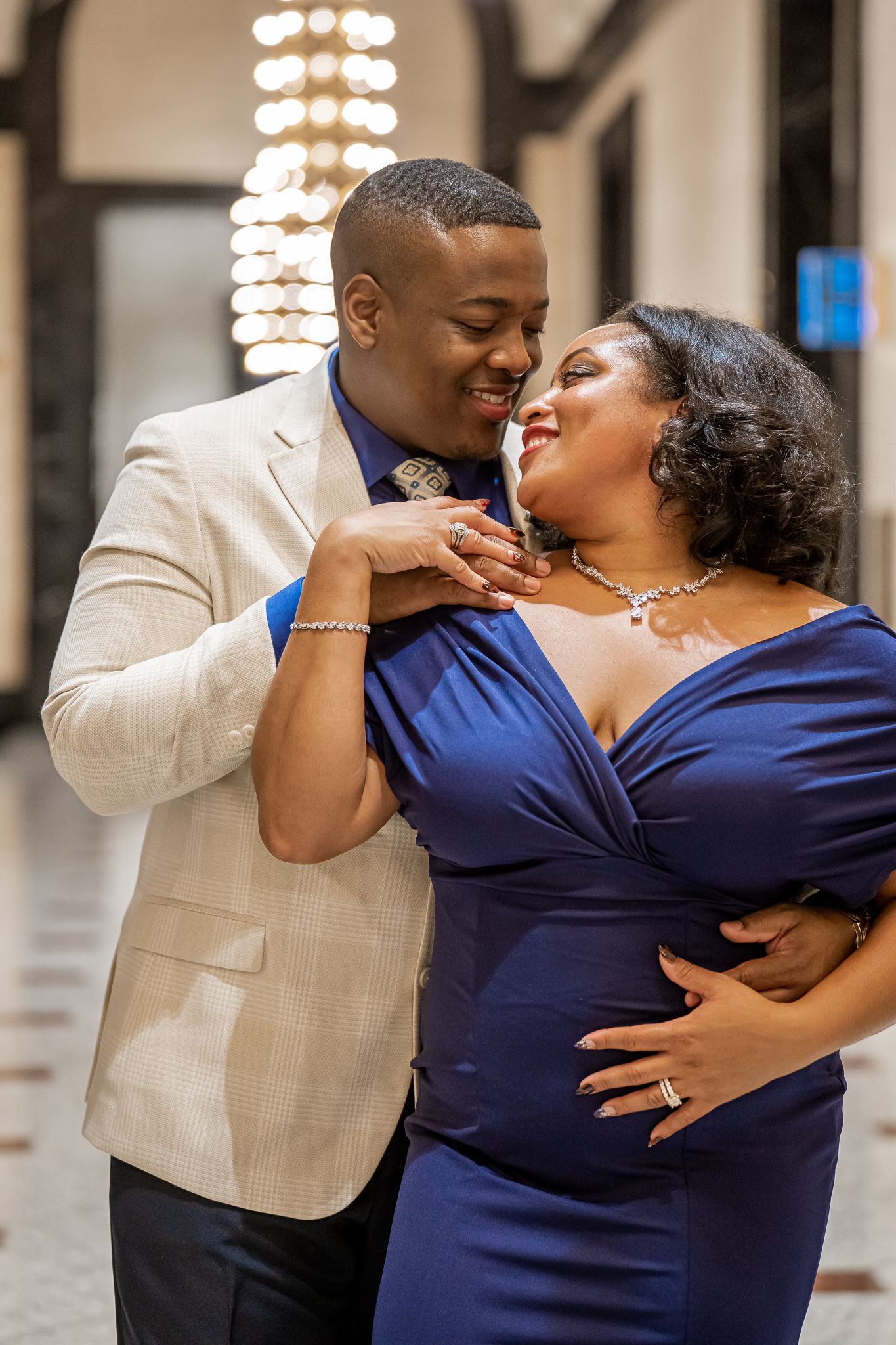 Couple shares tender moment in chandelier-lit hall, woman in blue gown displays engagement ring.