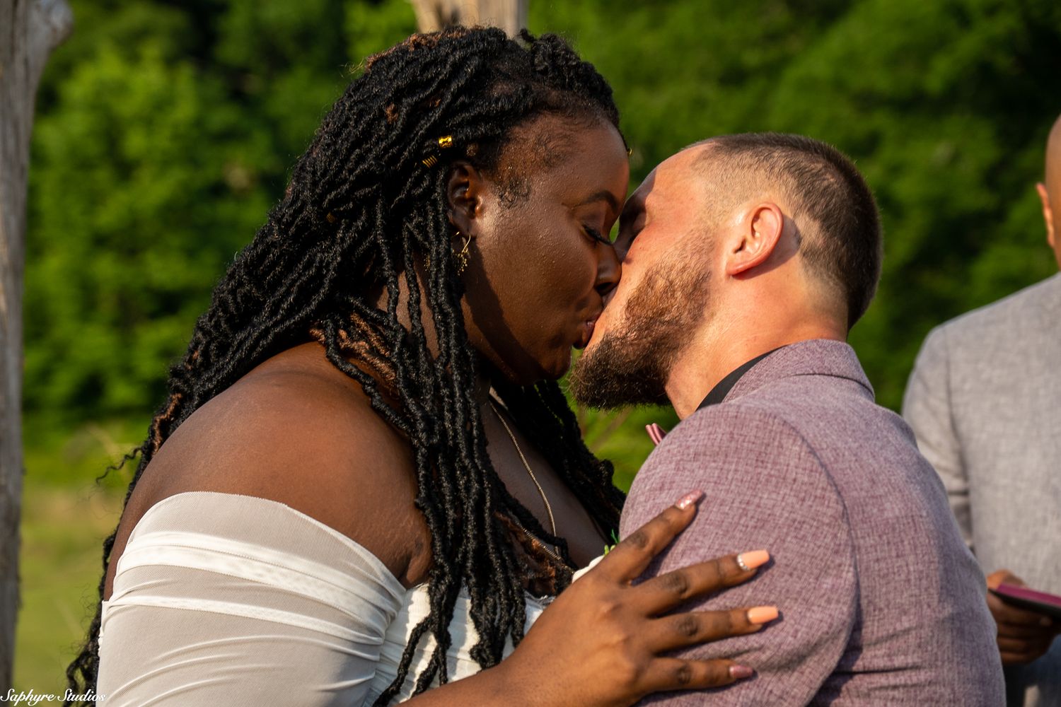 Interracial couple shares first kiss as newlyweds during outdoor wedding ceremony surrounded by lush greenery.