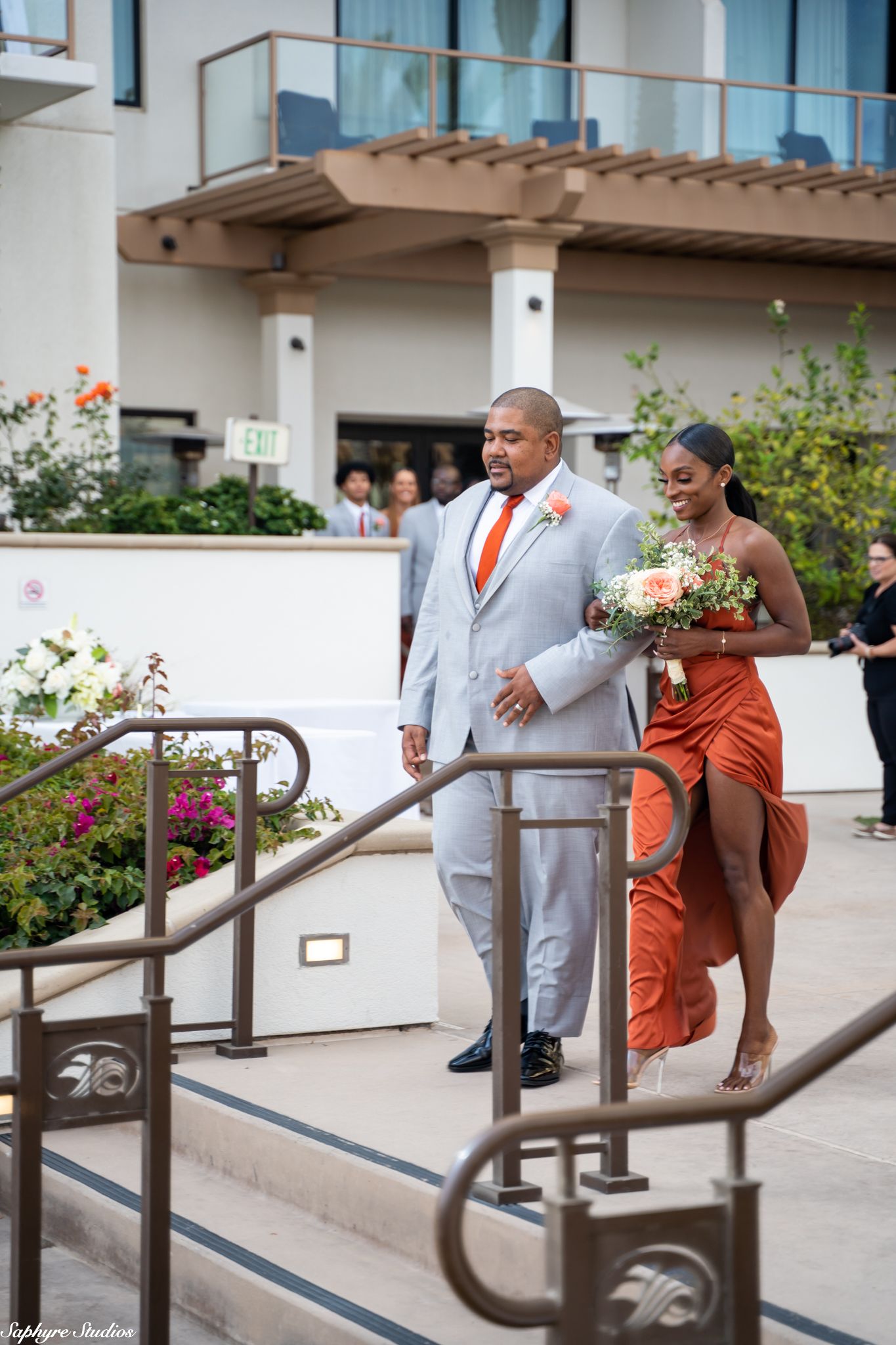 Groomsman in gray suit with orange tie escorts bridesmaid in rust orange dress at outdoor wedding.