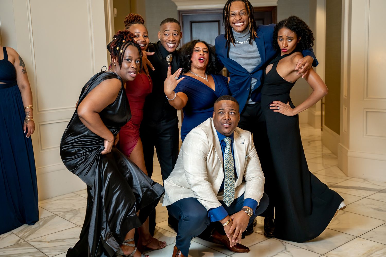 Lively group of eight in formal wear posing with fun expressions in an elegant hallway.